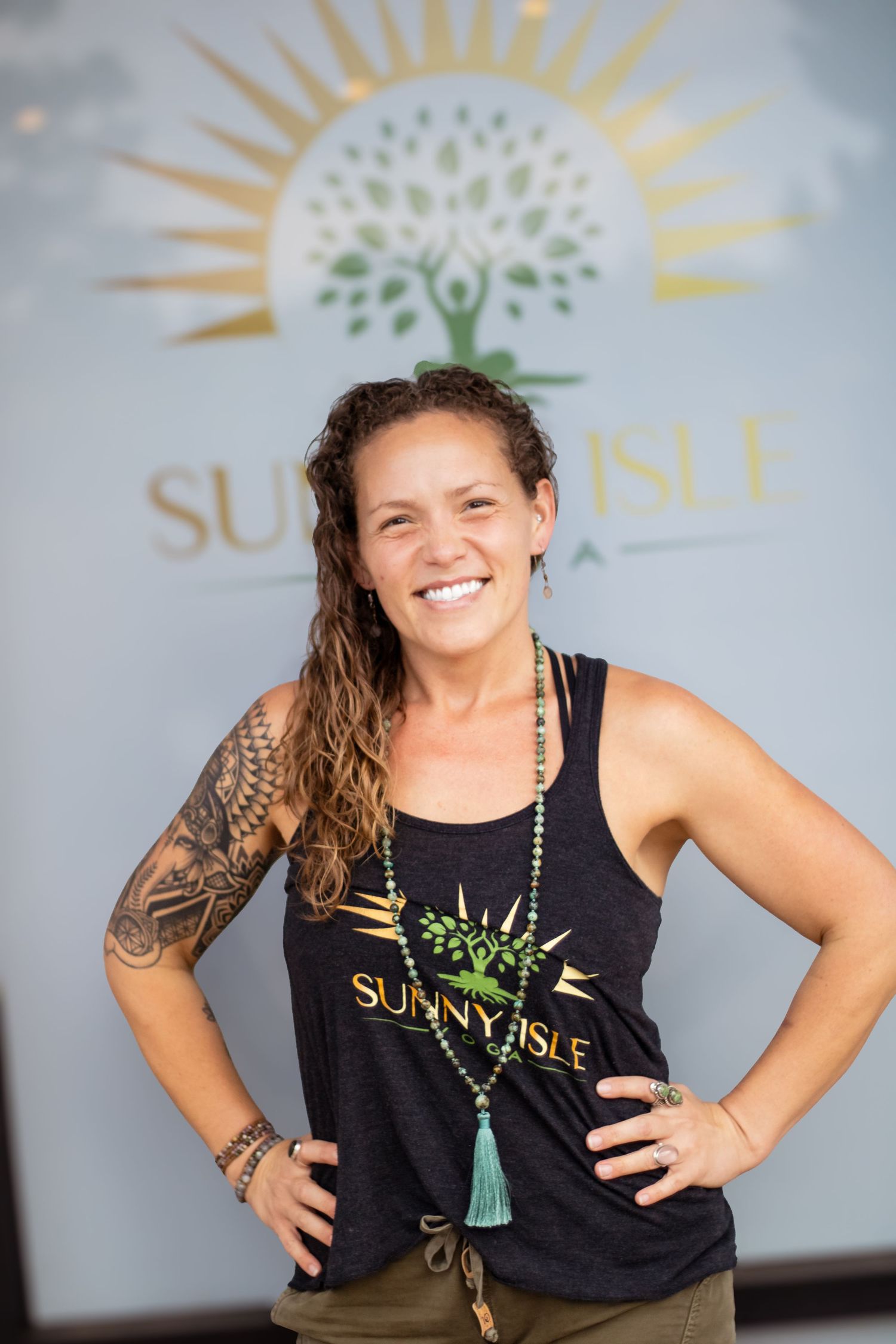 A person in a black tank top with tattoos poses against a gray backdrop with a sunburst logo.