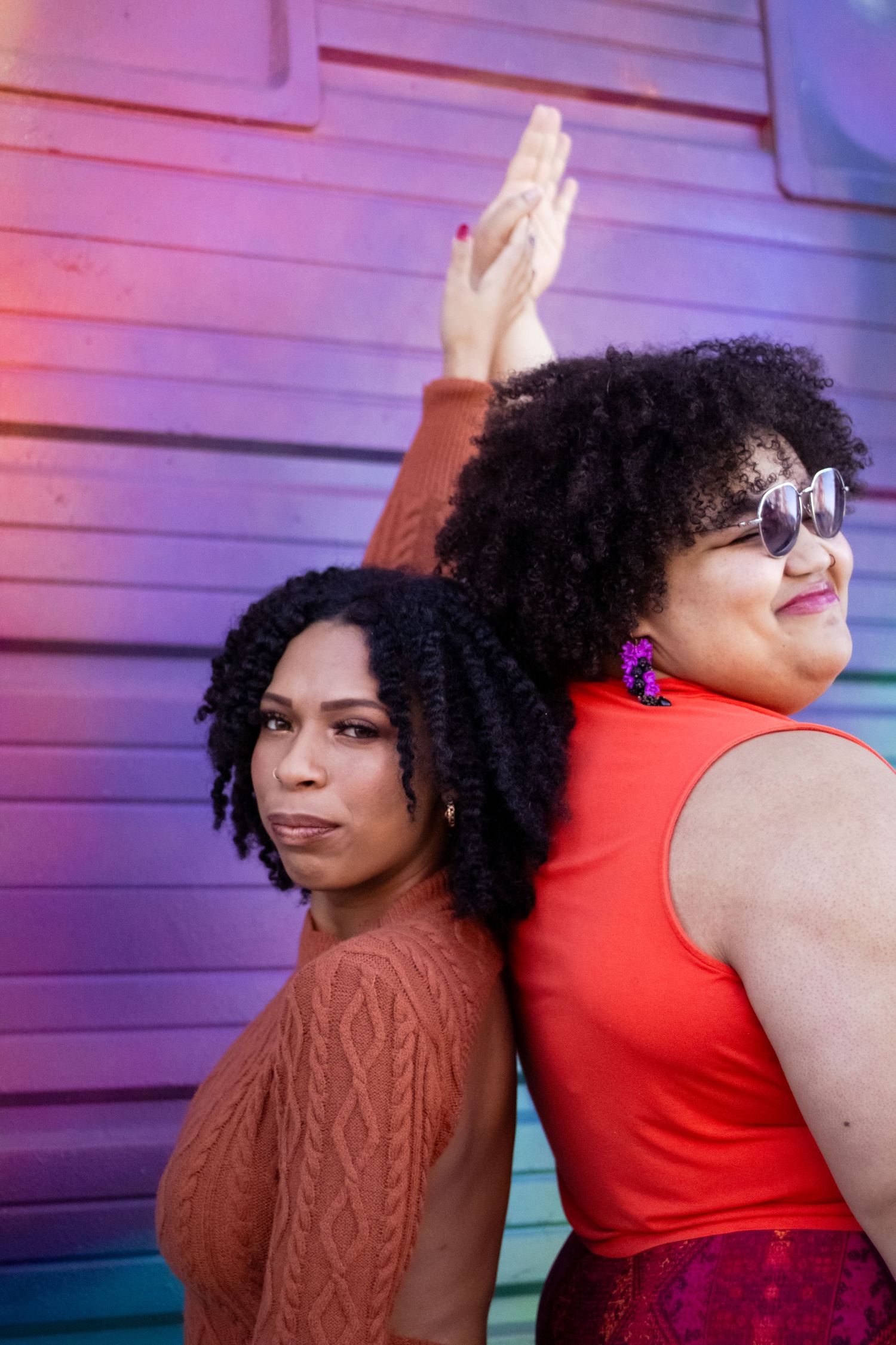 Two women in red and brown outfits dance together against a pink-lit wall in a dynamic pose.