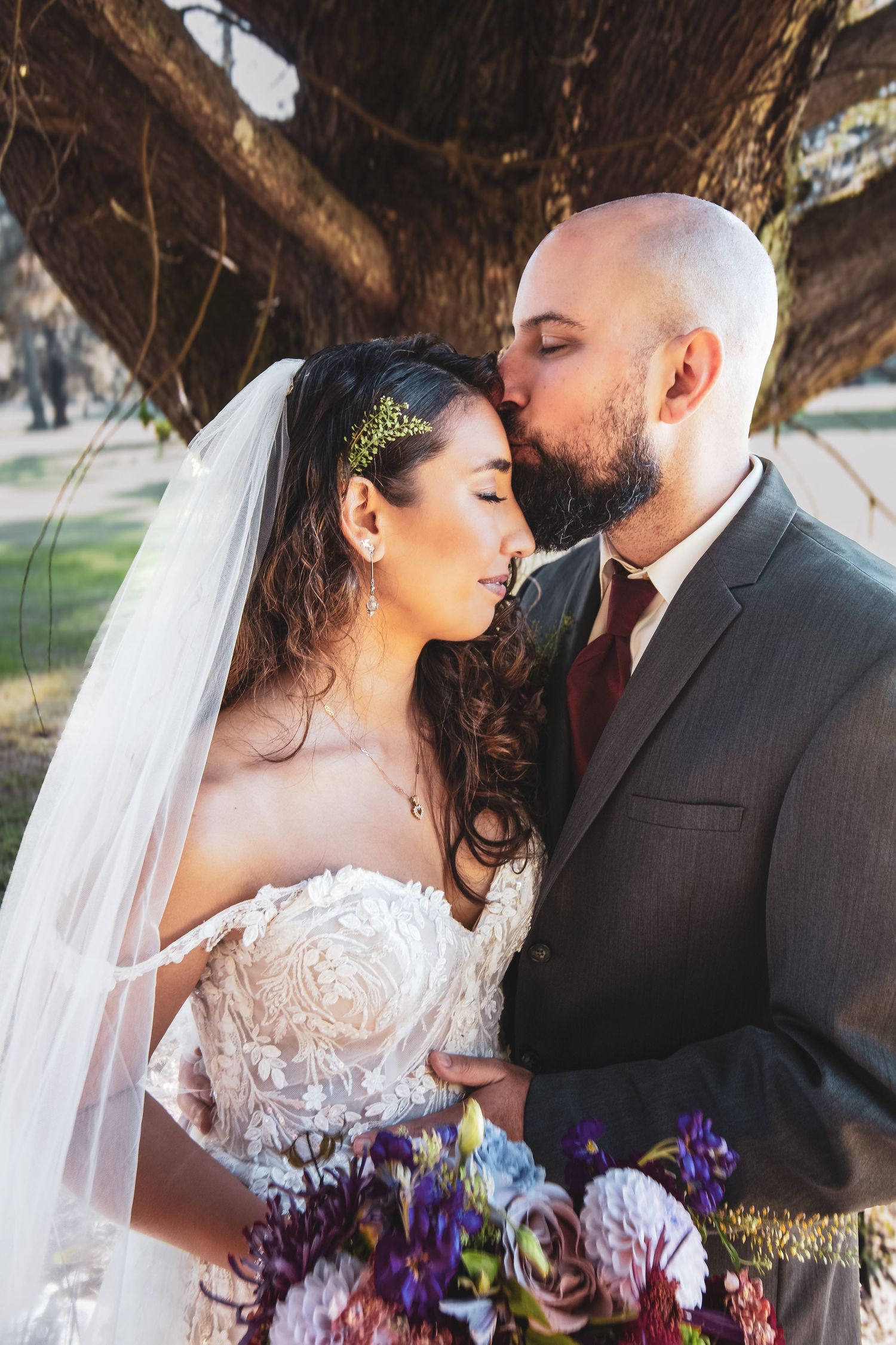 A tender wedding moment captured under a tree, with a stunning bouquet of purple and blue flowers.