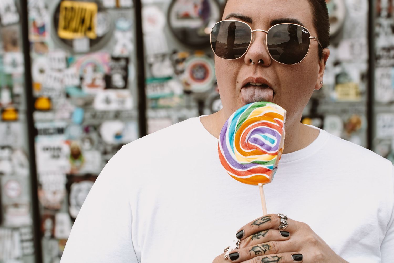 Two people in white shirts enjoy lollipops while standing in front of a wall covered with stickers and posters.