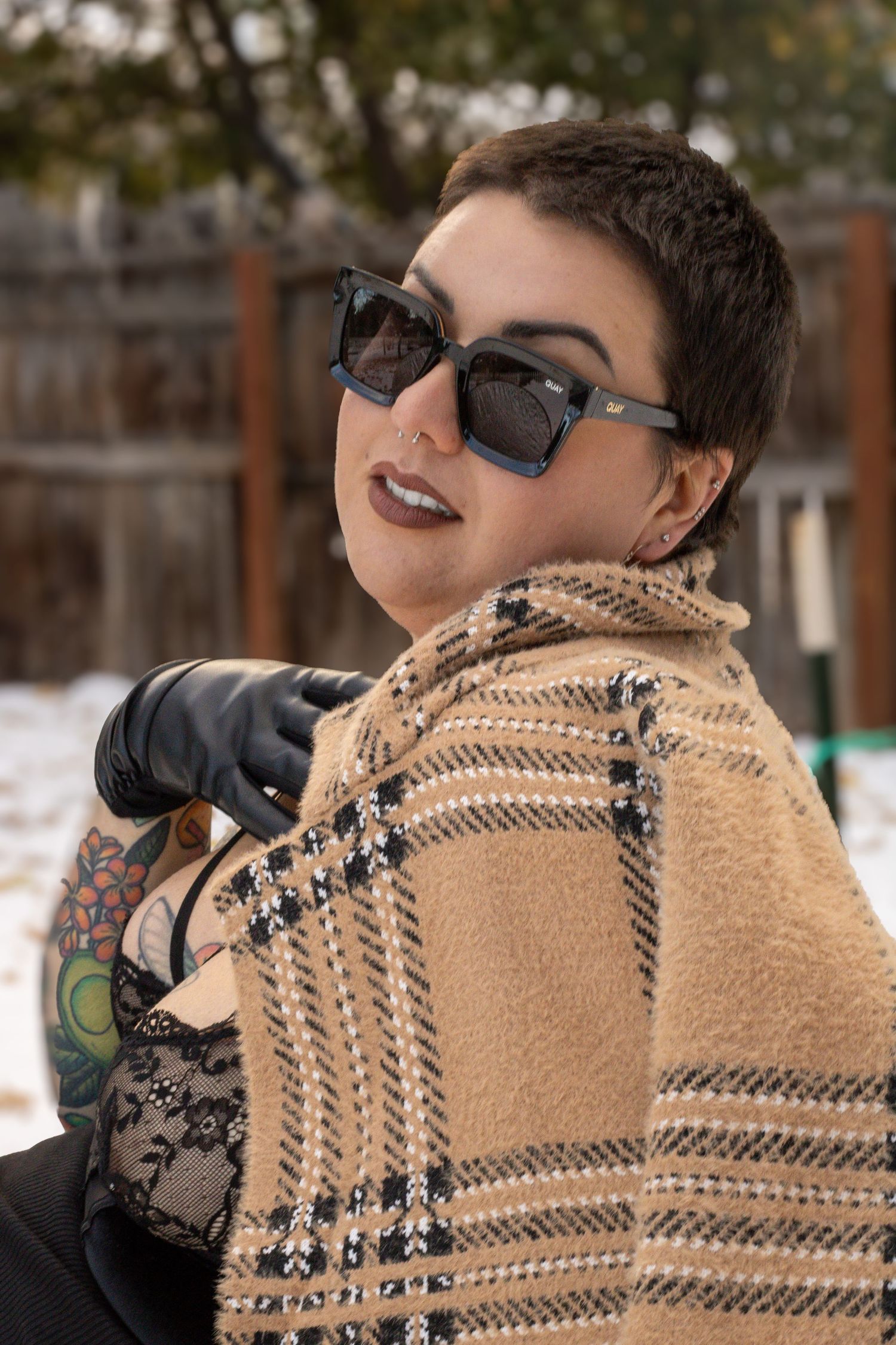 A person in sunglasses and plaid blanket wrap poses outdoors in winter with short dark hair and bold lipstick.