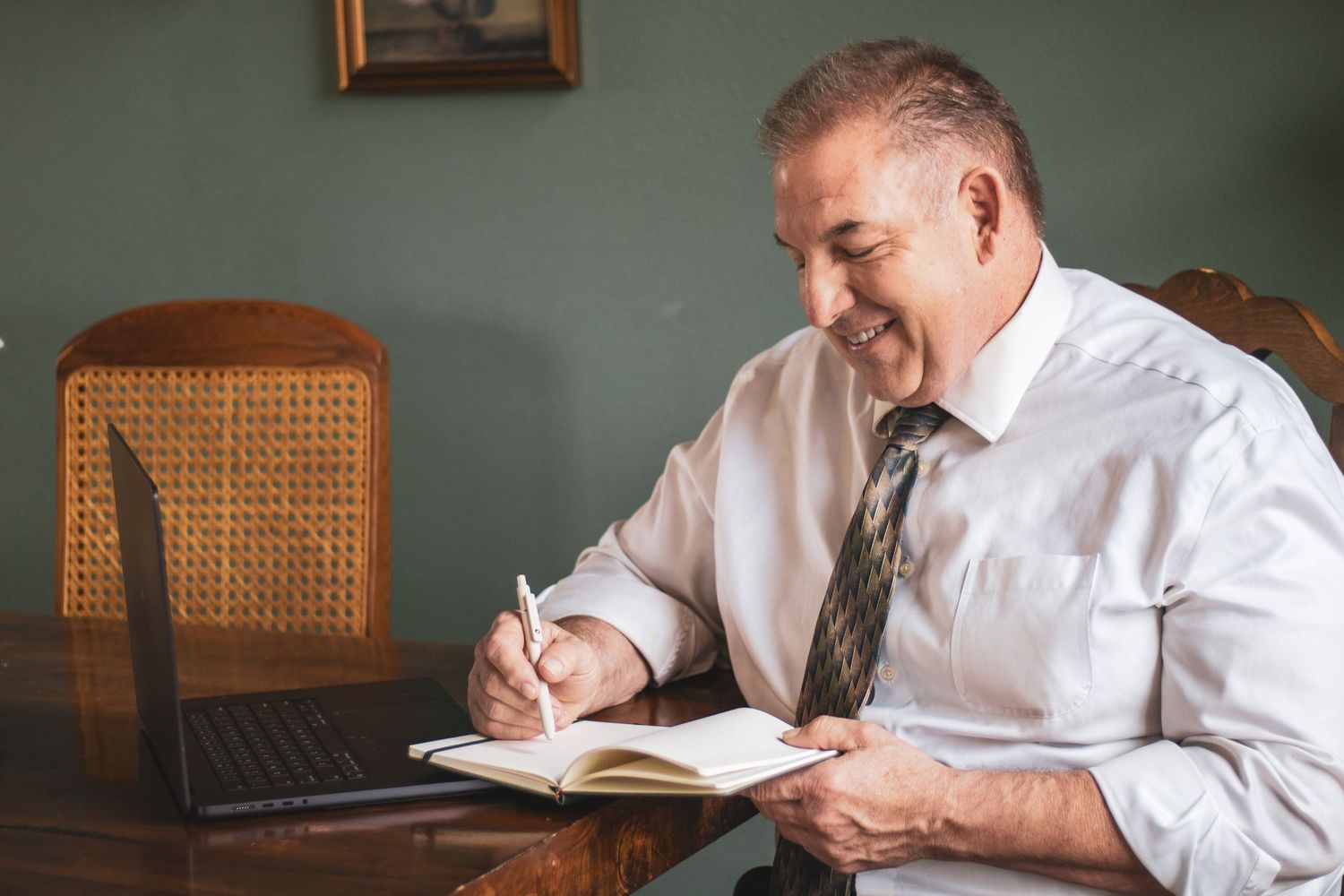 Professional in white shirt and tie writes in notebook while sitting at wooden desk with cane back chair.