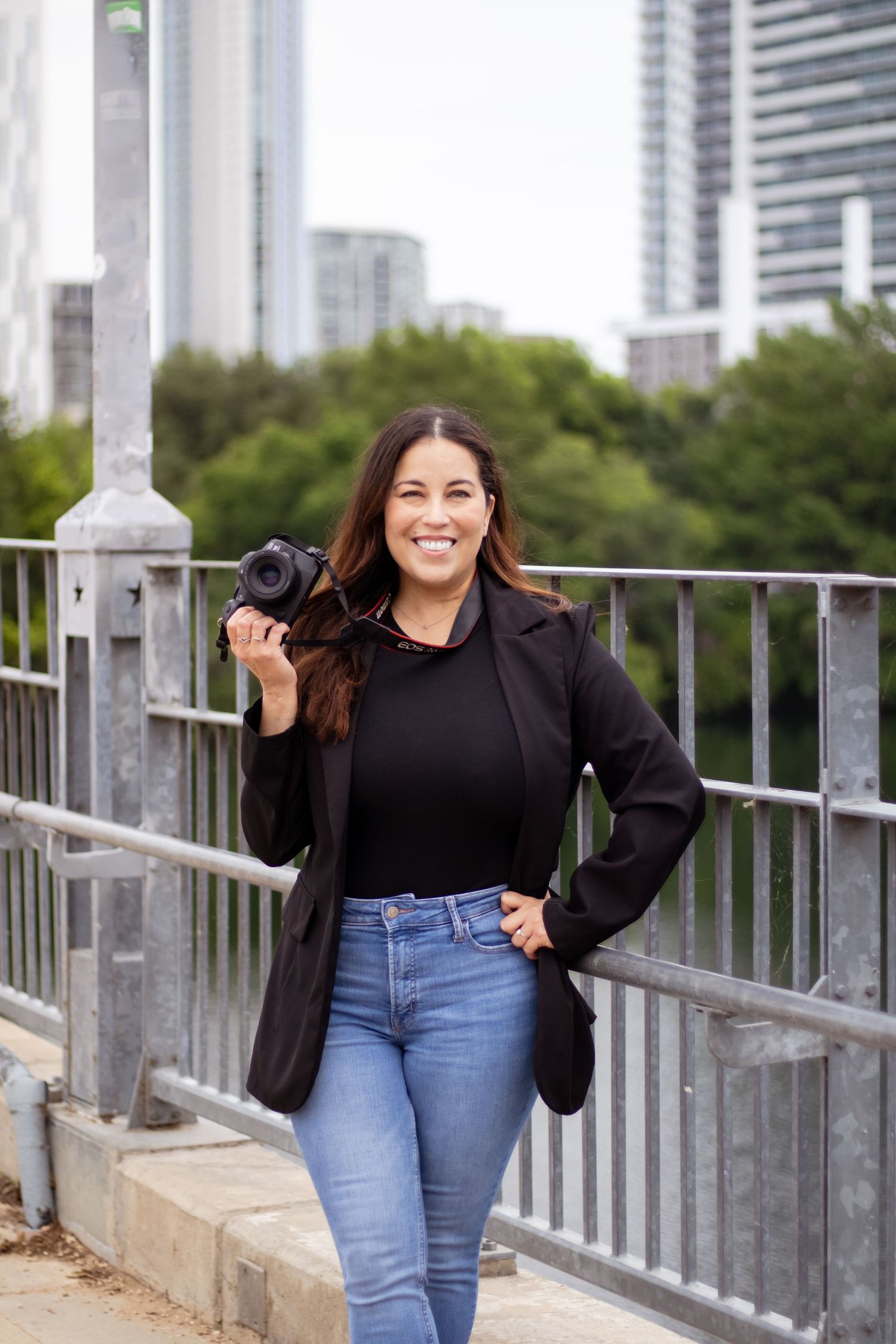 Woman in black top and blue jeans poses casually against railing with city skyline and trees in background.
