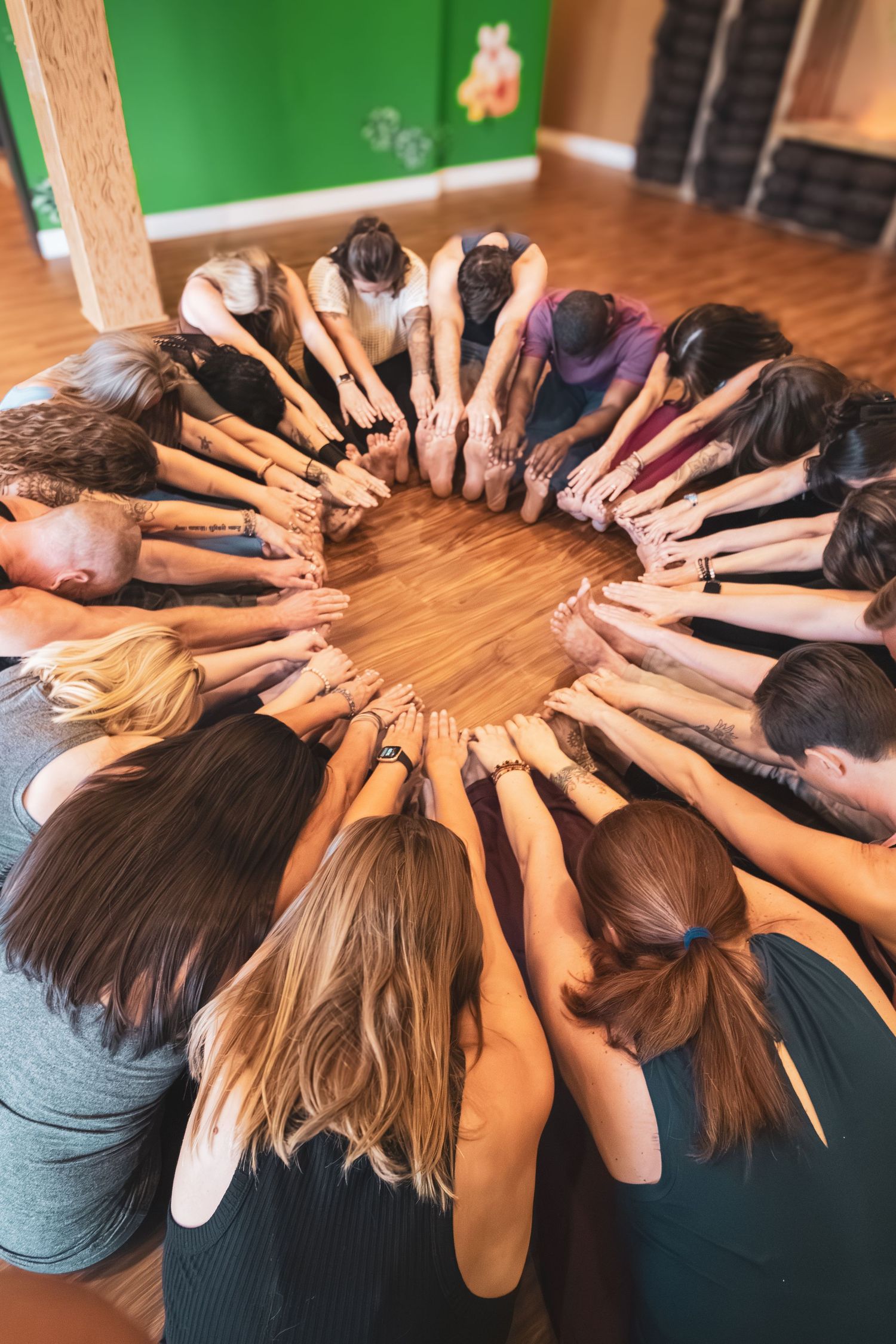 A group of people form a circle with their arms extended inward, creating a unity huddle on a wooden floor.
