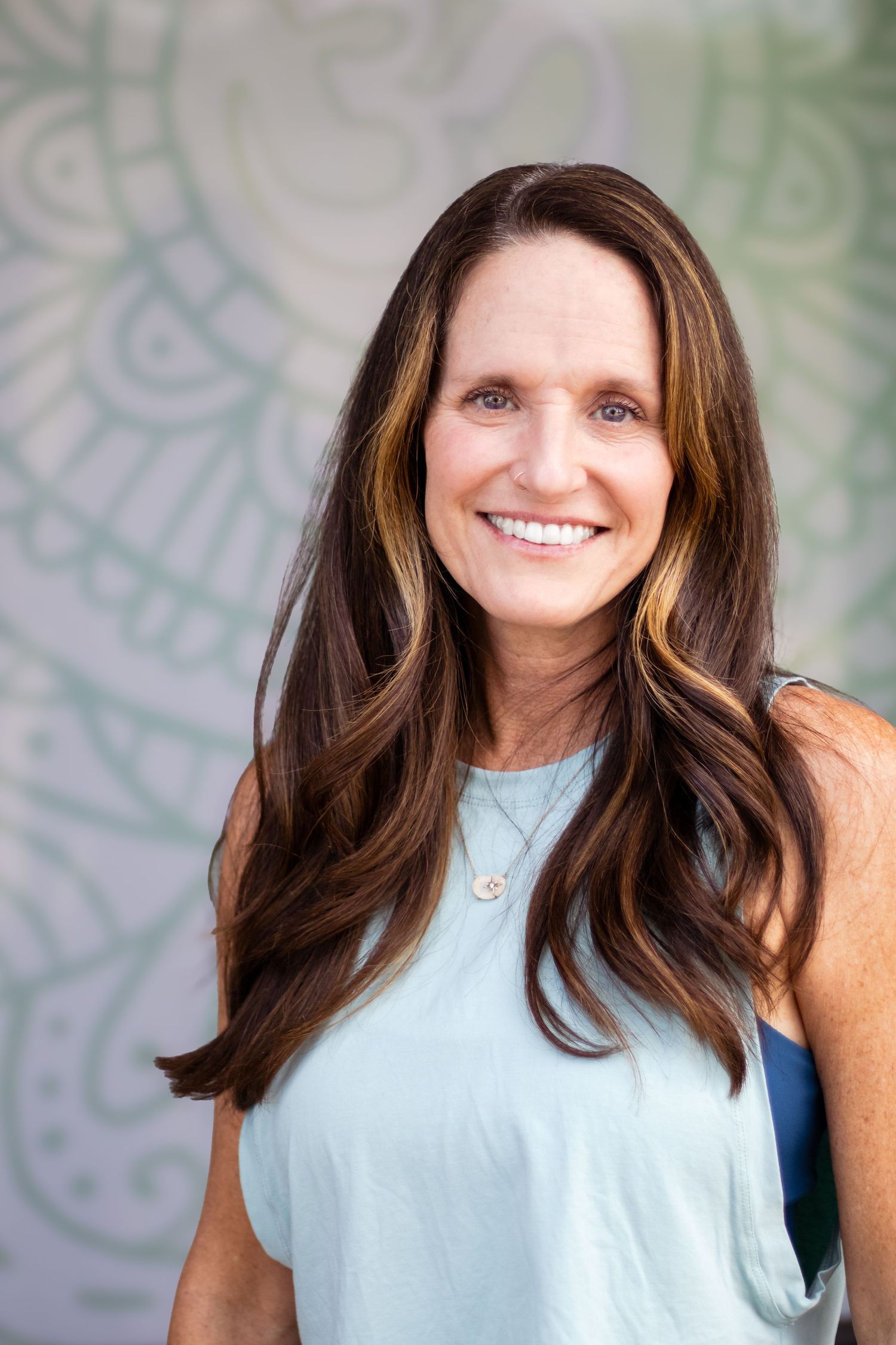 Woman in light blue sleeveless top smiling against a pale patterned background.
