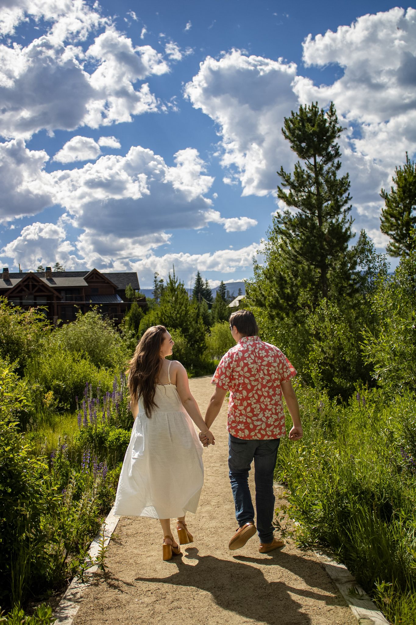 A couple walking hand in hand along a sunny dirt path surrounded by evergreen trees and lush vegetation on a cloudy day.