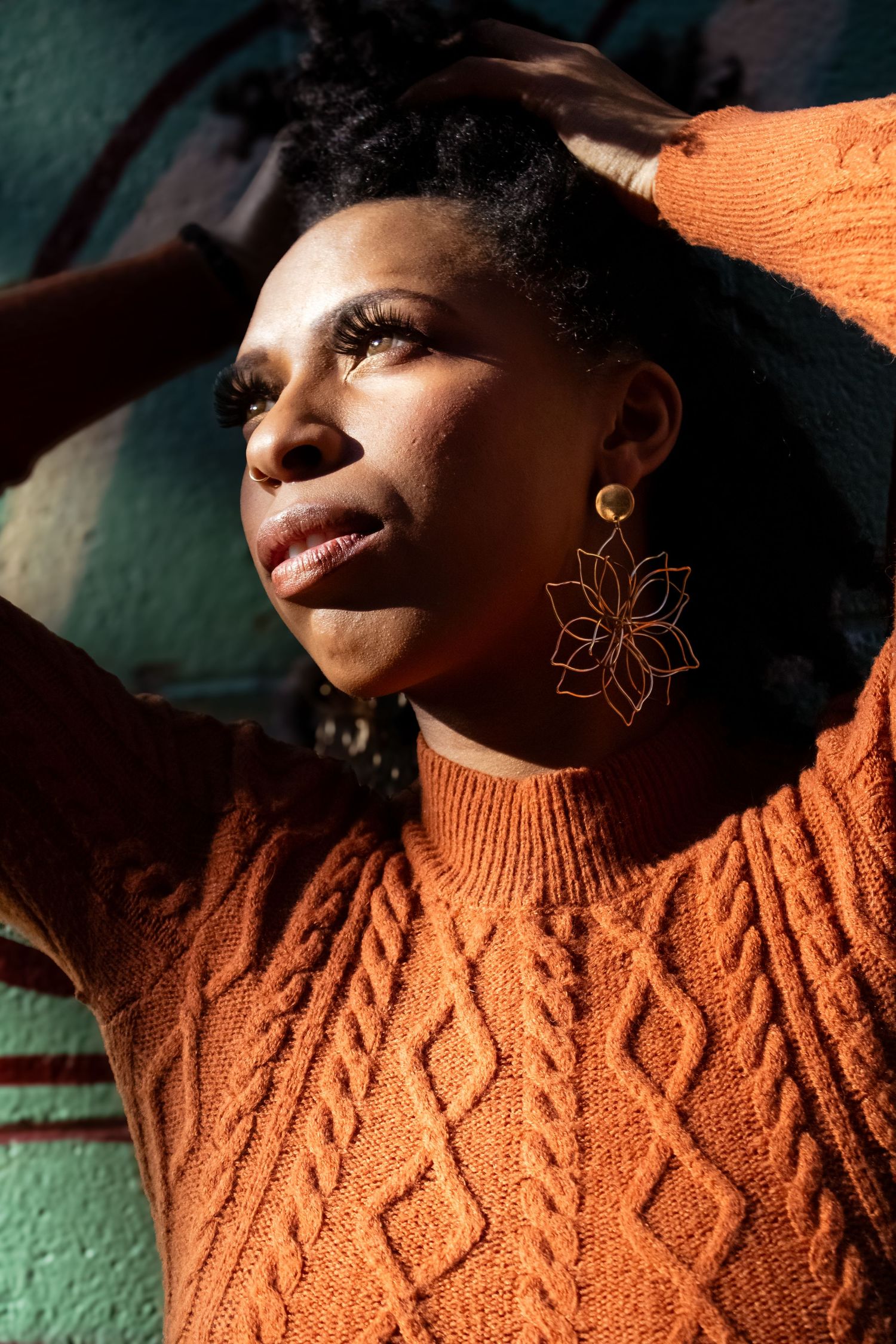 Person wearing an orange cable knit sweater and floral earring poses in dramatic side lighting against a green wall.