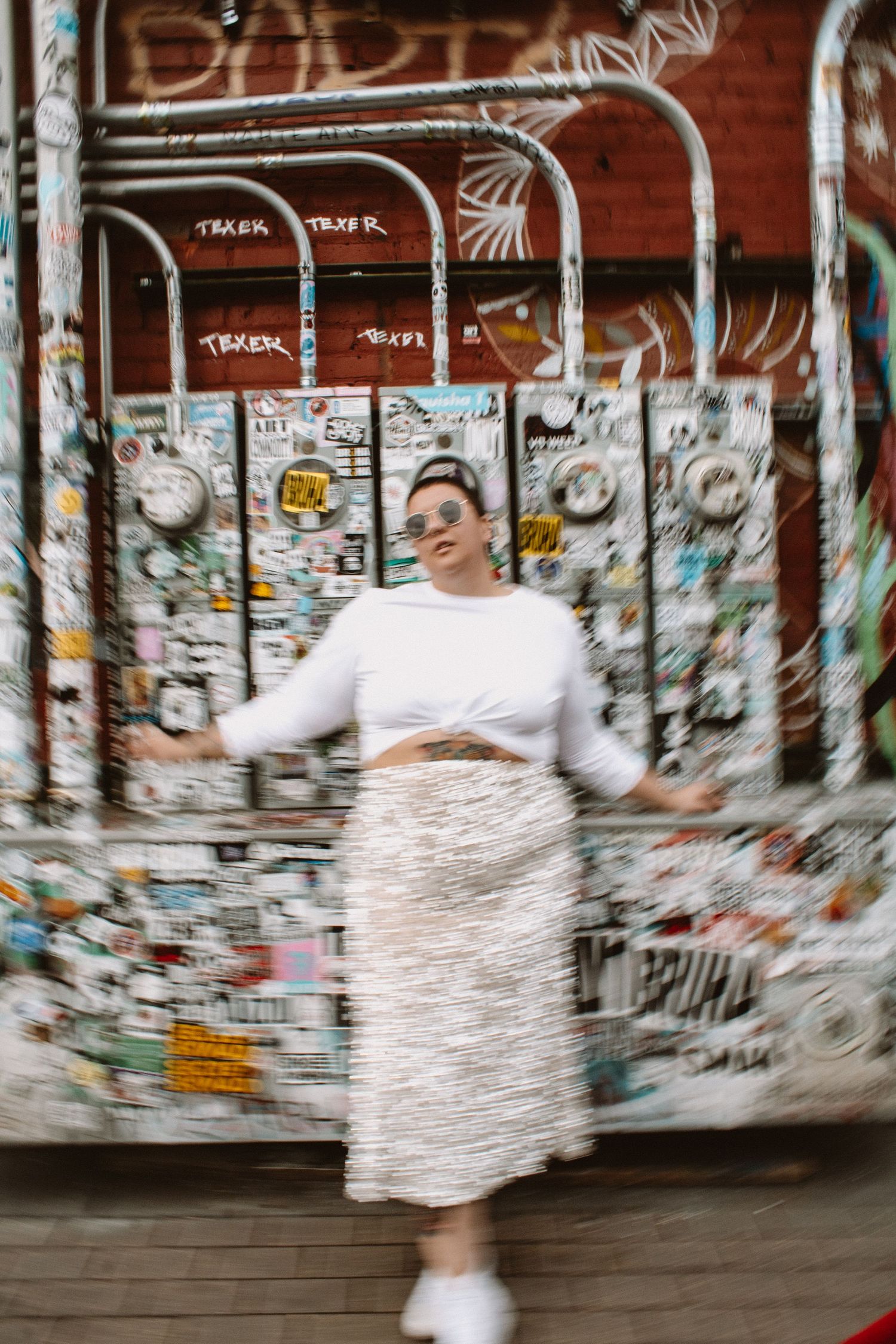Motion-blurred figure in white dress spins against graffiti-covered wall creating ethereal movement effect in urban setting.