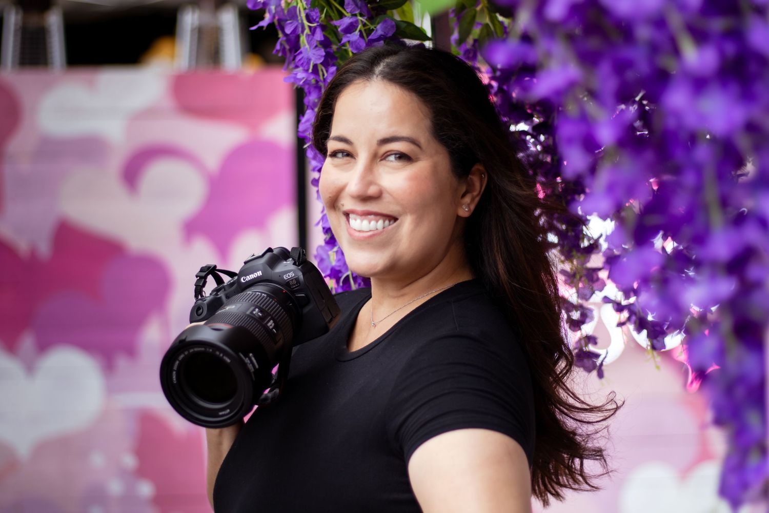 Professional photographer with long dark hair holding a DSLR camera stands in front of vibrant purple flowers.