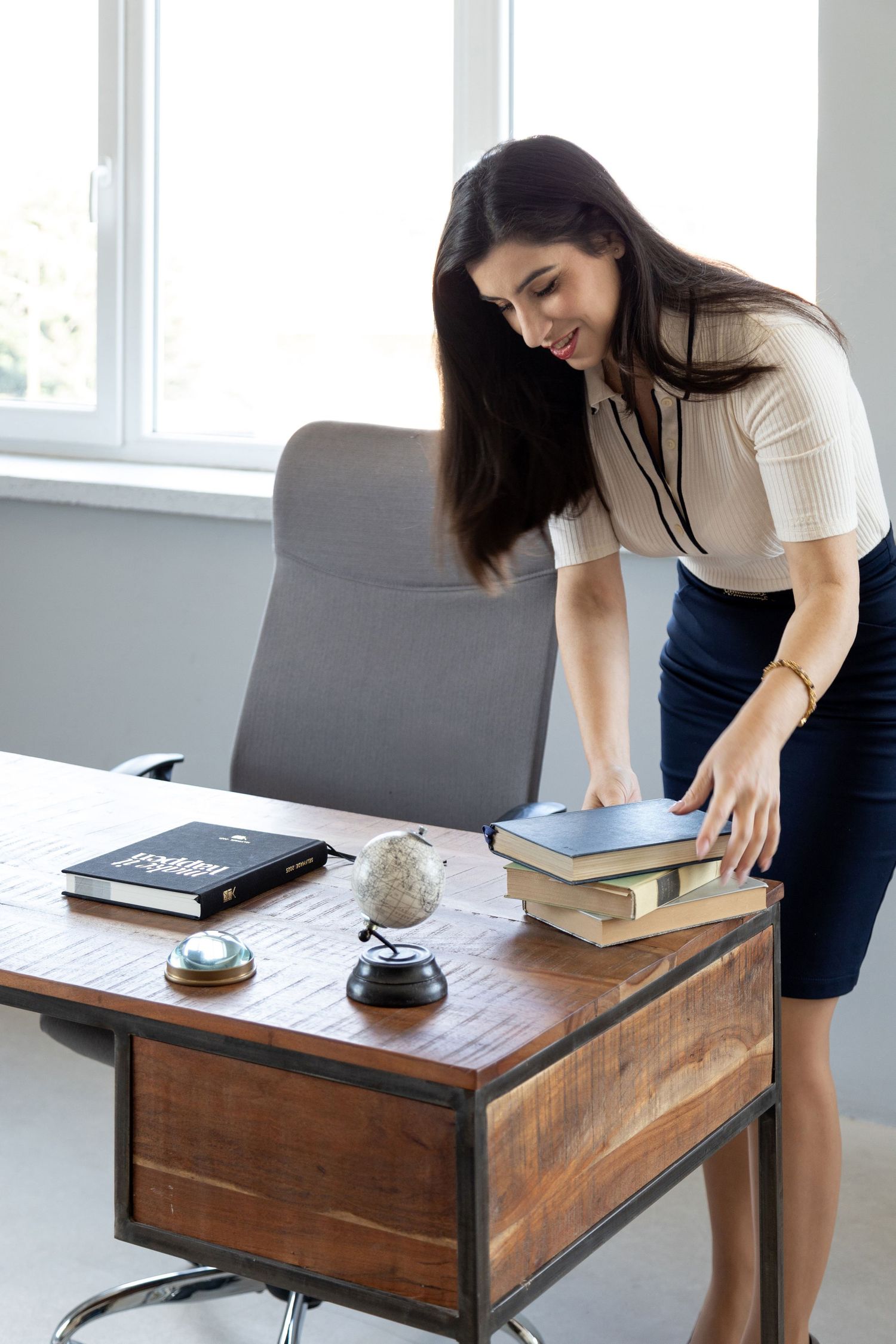 A young businesswoman in a white blouse and navy skirt arranges books on a wooden office desk with a globe and bell.