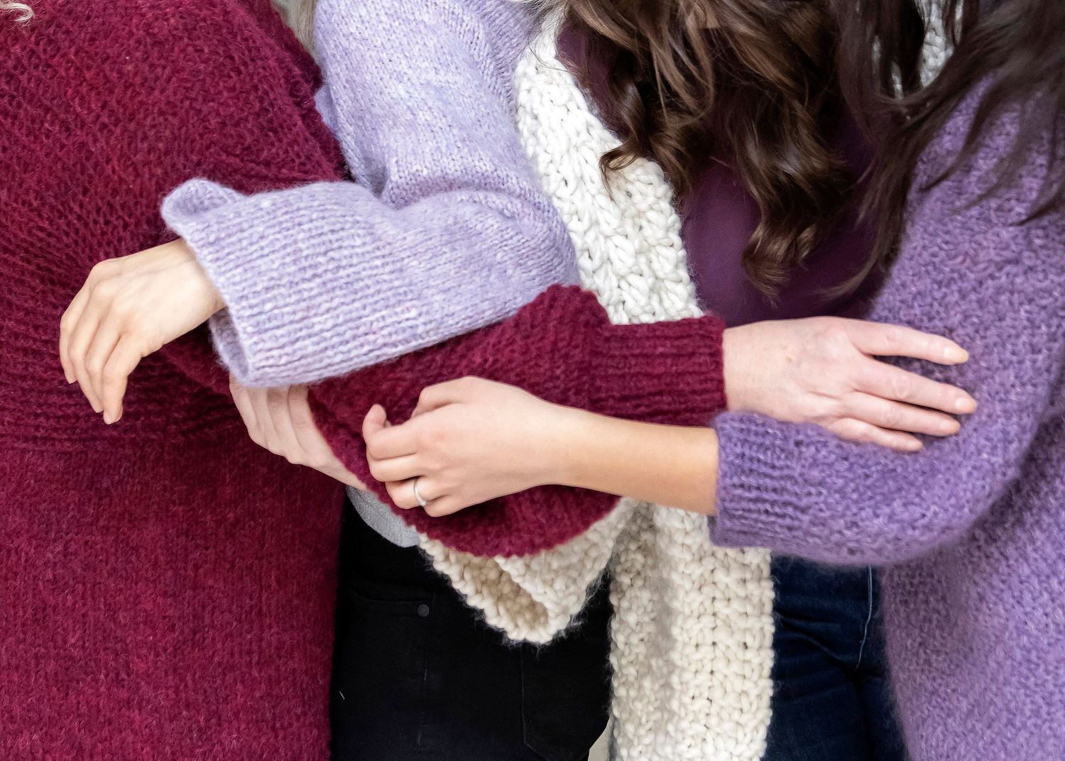 Three people in handknit sweaters in burgundy, lavender, and purple linking arms closely.