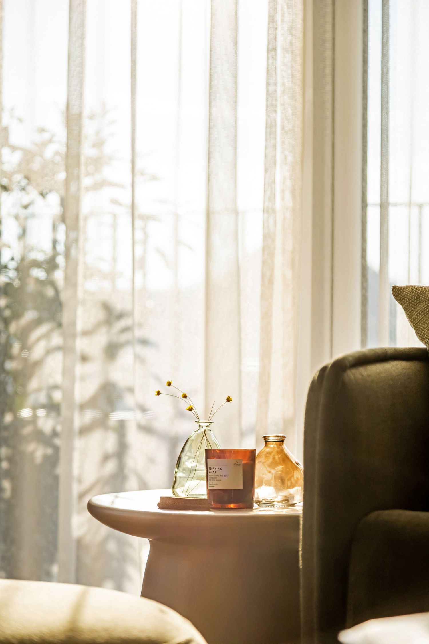 Cozy sunlit reading nook with sheer curtains, a candle, small vase with dried flowers, and amber glass bottle on a round side table.