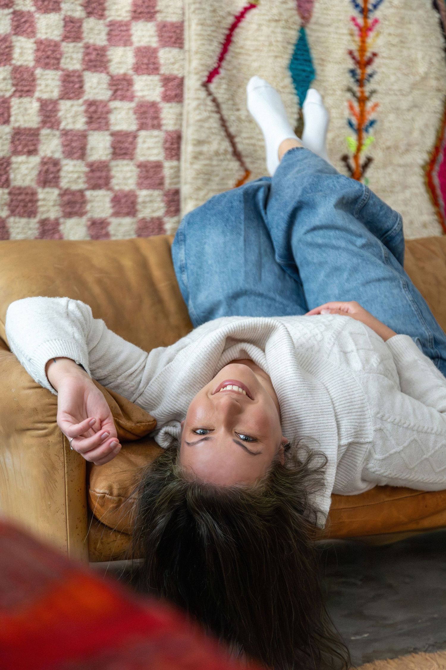 Woman in jeans and white sweater lying upside down on a leather sofa, smiling, with colorful rugs in the background.