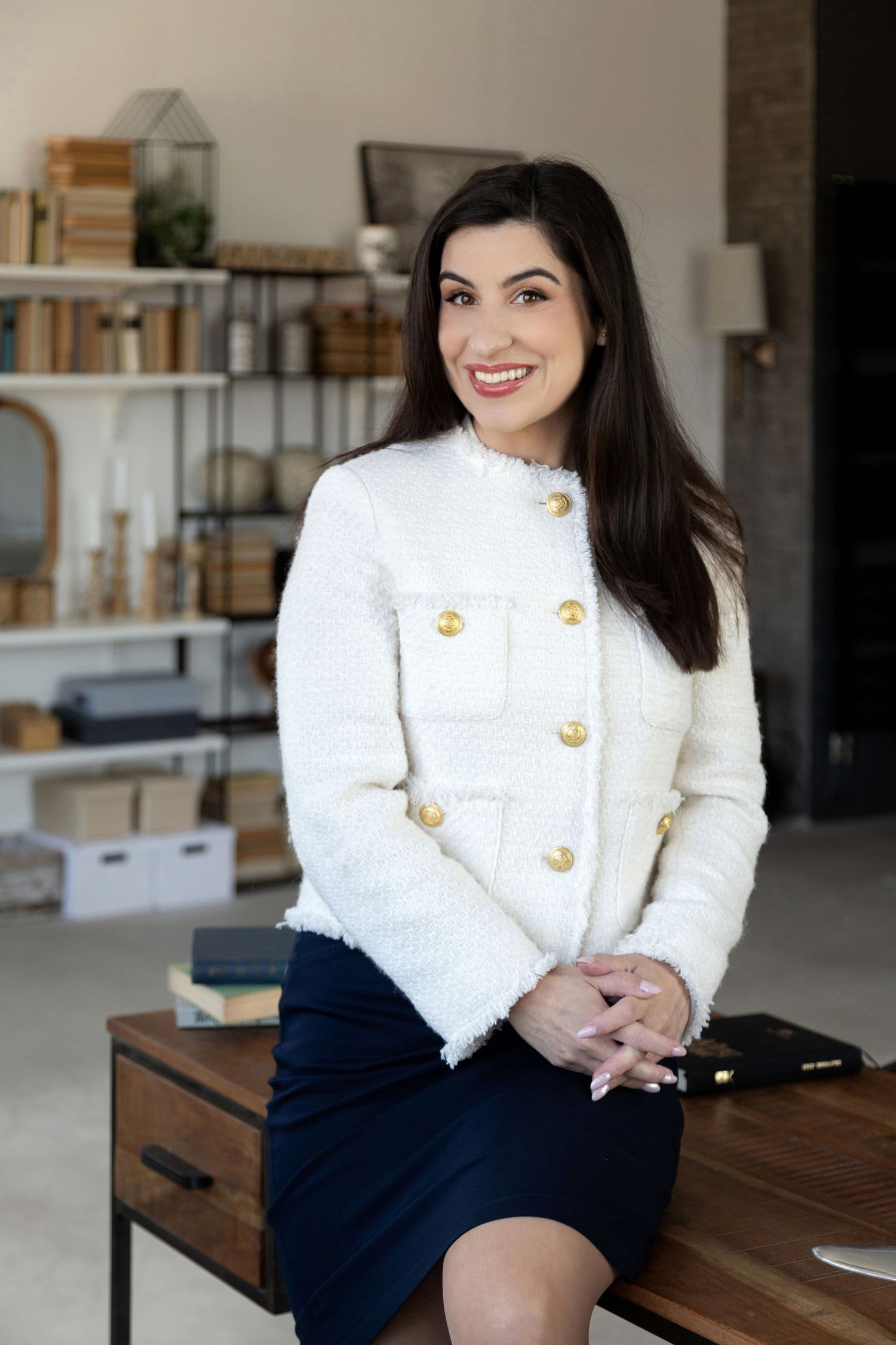 Professional woman in white tweed jacket and navy skirt sitting on desk in modern office setting.