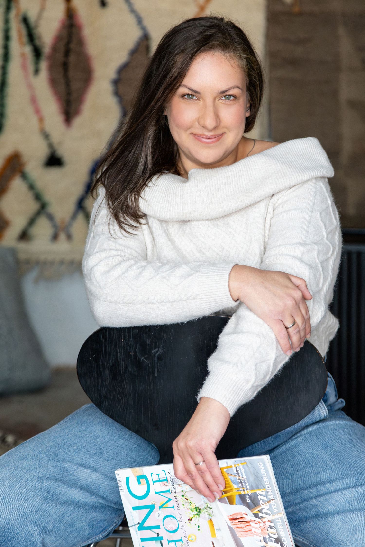 Smiling brunette woman in white off-shoulder sweater sitting cross-legged holding a home magazine.