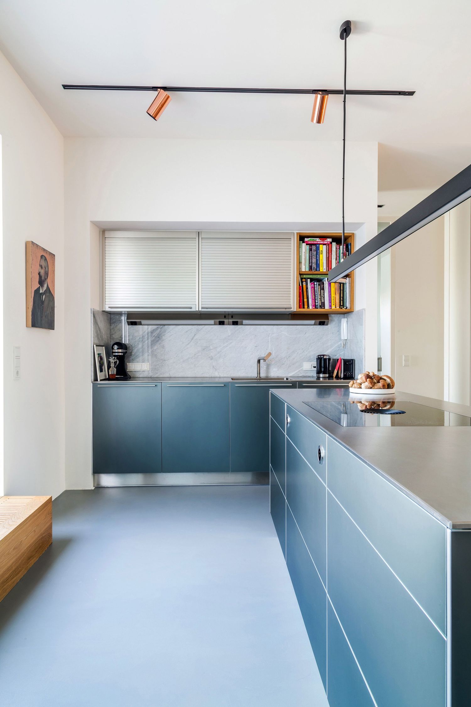 Sleek modern kitchen with blue-gray cabinetry, marble backsplash, track lighting, and colorful bookshelf accent.