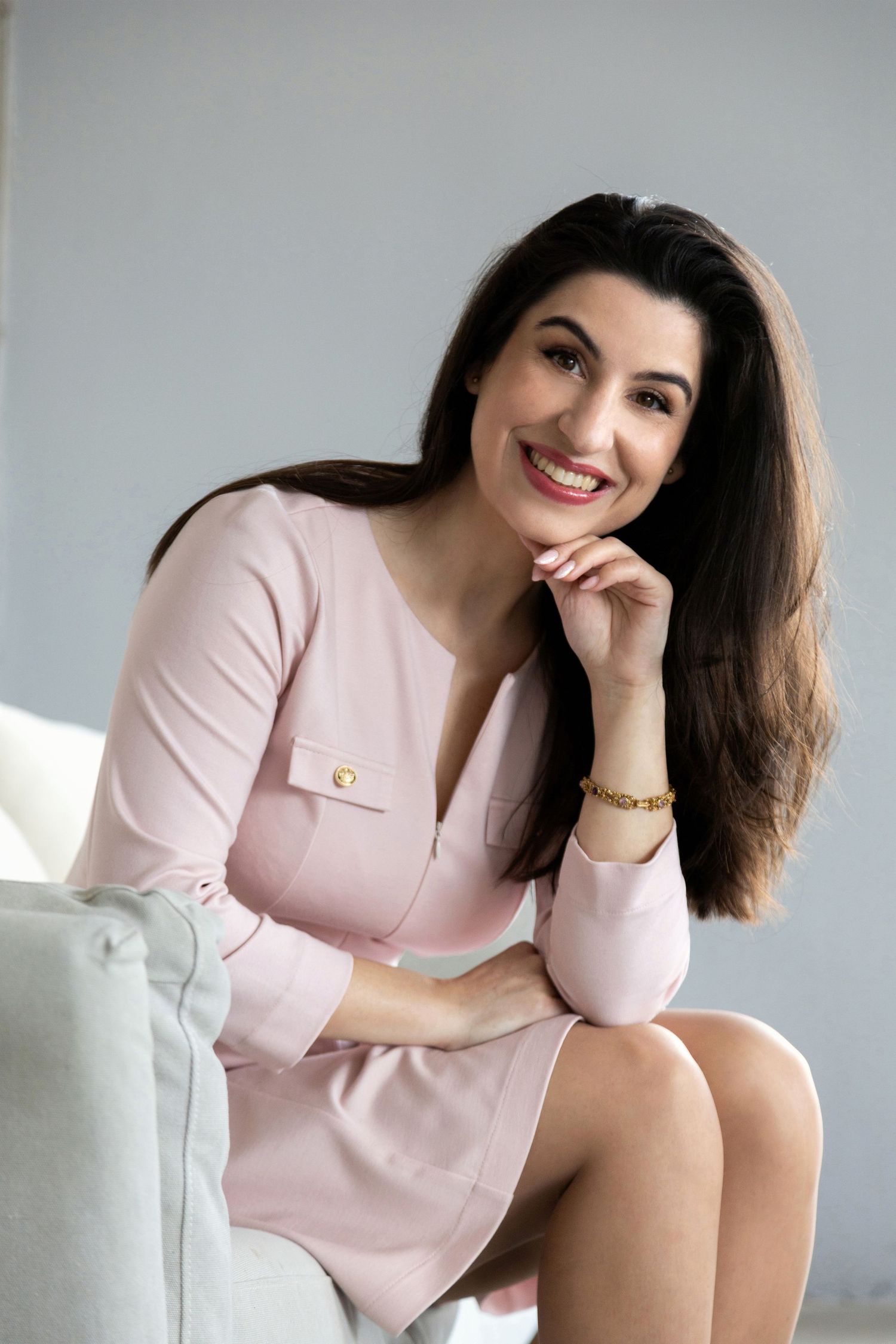 Smiling brunette woman in pink dress sitting elegantly in a chair with gold bracelet and confident pose.