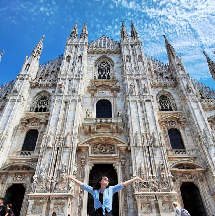 Person standing with arms outstretched before the ornate Gothic facade of Milan Cathedral, Duomo di Milano, Italy.