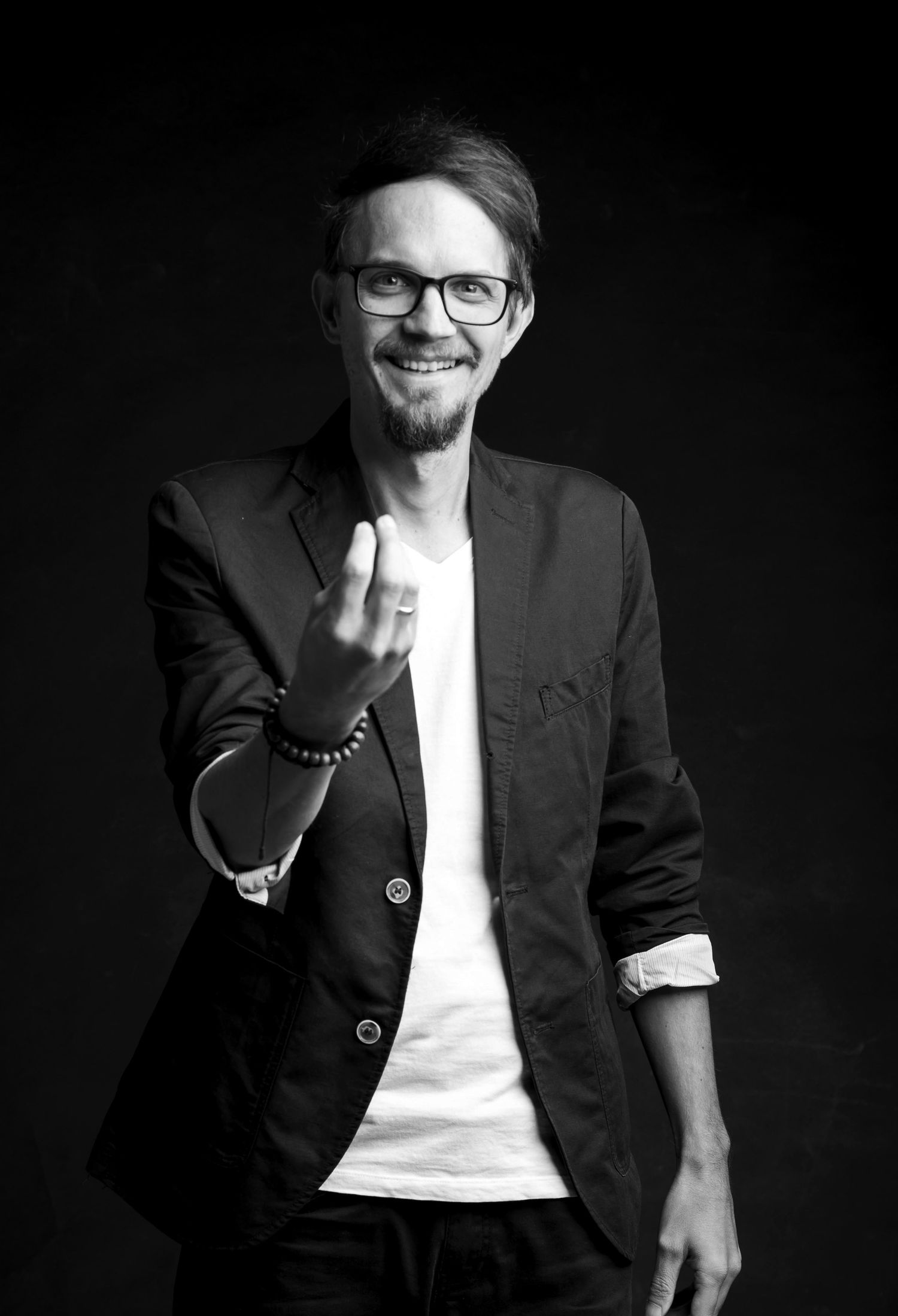 Black and white studio shot of smiling man in blazer and glasses making a beckoning hand gesture.