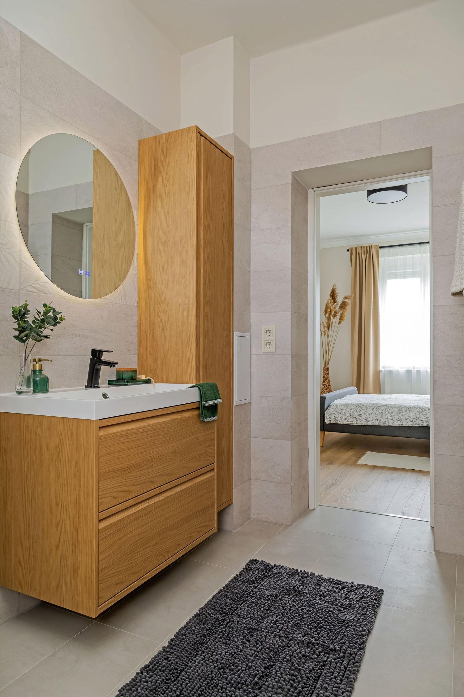 Contemporary bathroom with oak vanity, round backlit mirror, beige tiles, and doorway leading to bedroom.