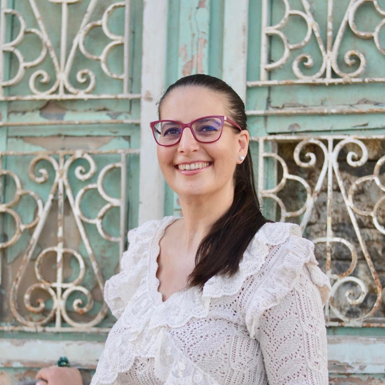 Smiling woman with red glasses and white lace top posing in front of ornate vintage mint green iron gate.