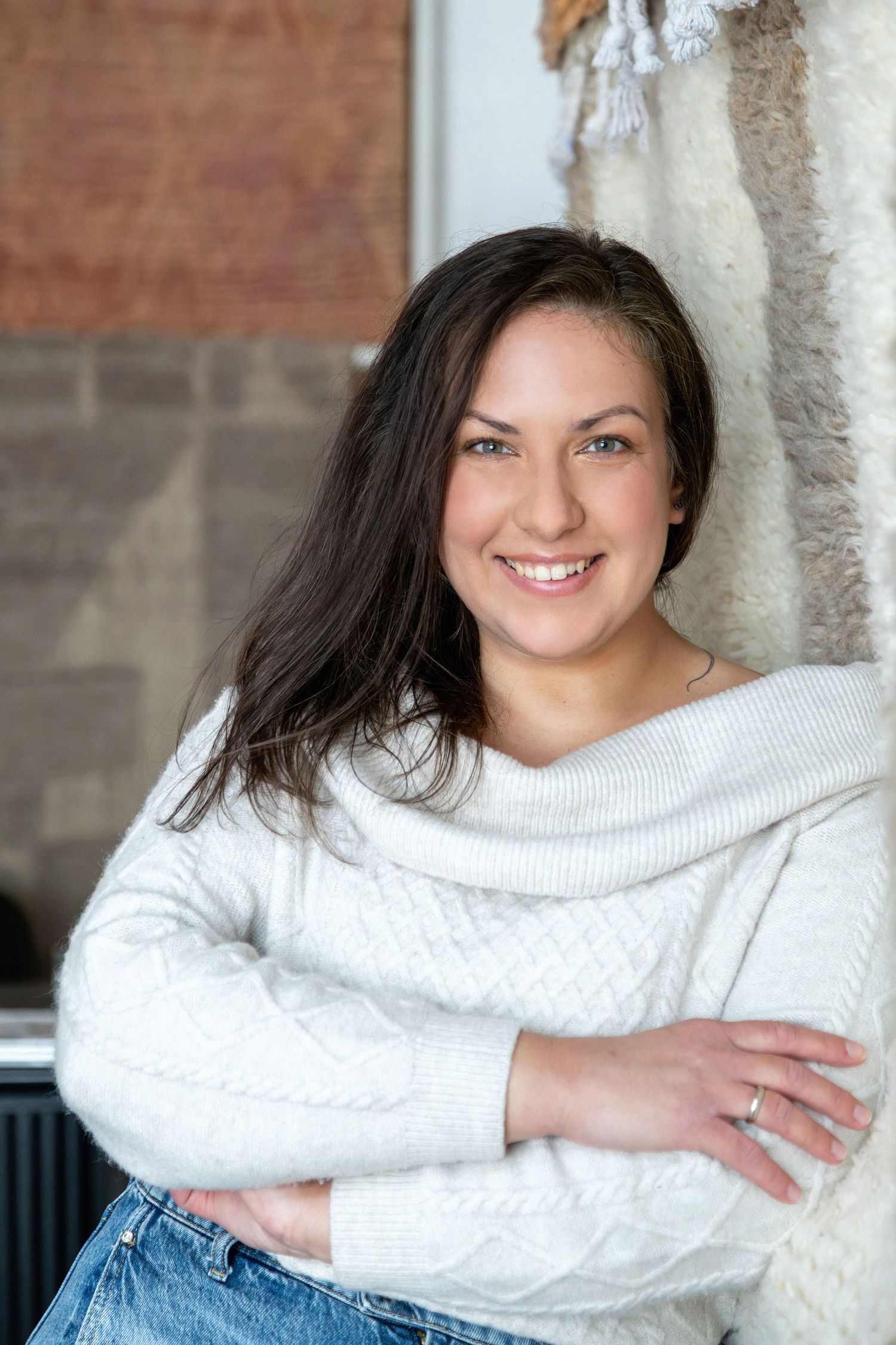 Smiling brunette woman in white cable-knit sweater with arms crossed, leaning against wall.