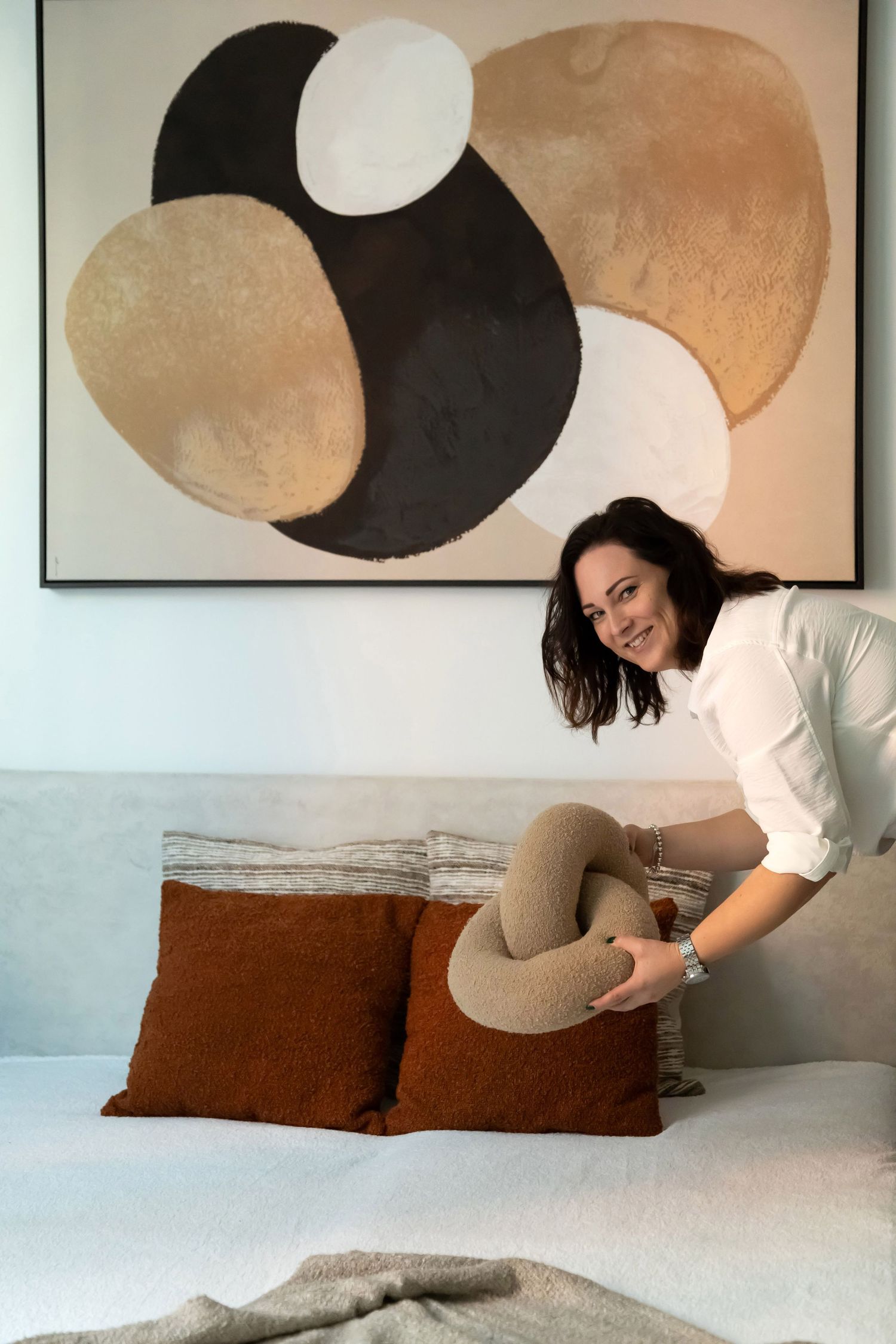 Happy woman in white shirt styling a bedroom by placing a textured knot cushion on a neutral-toned bed.