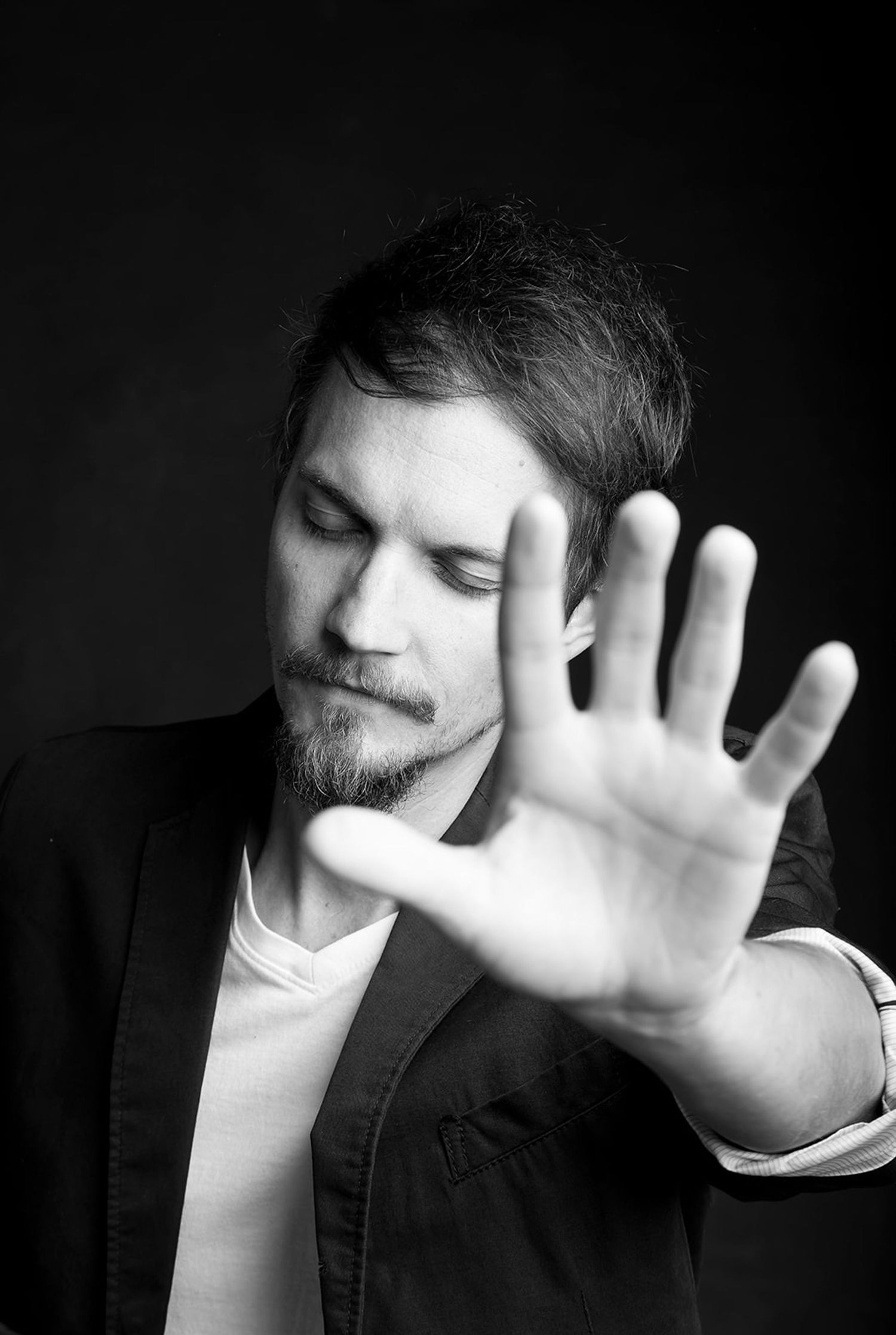 Black and white dramatic portrait of bearded man holding hand toward camera against dark background.