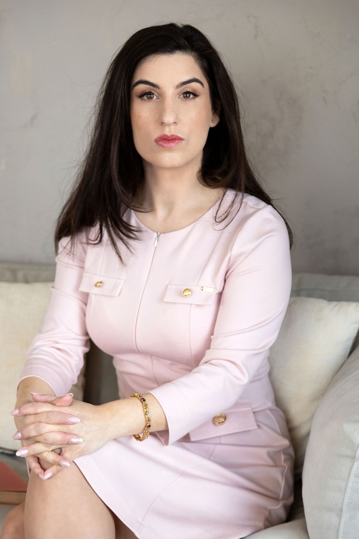 Professional woman in pink dress with gold buttons sits confidently, dark hair, neutral expression, indoor setting.