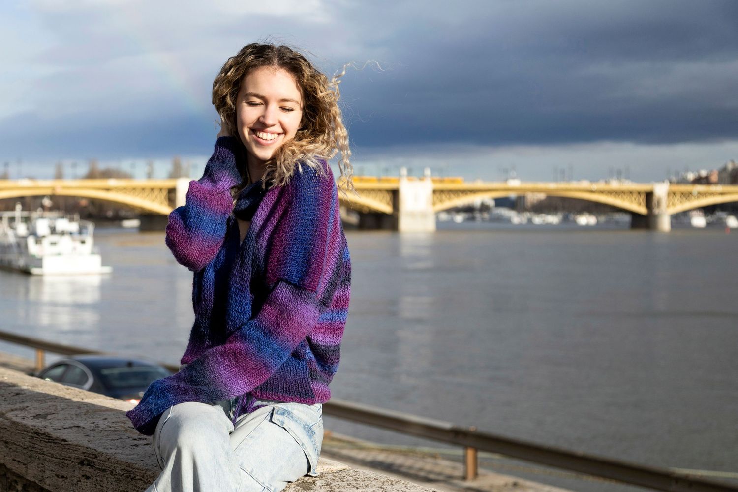 Happy woman in a colorful knit sweater smiling by a river bridge on a cloudy day.