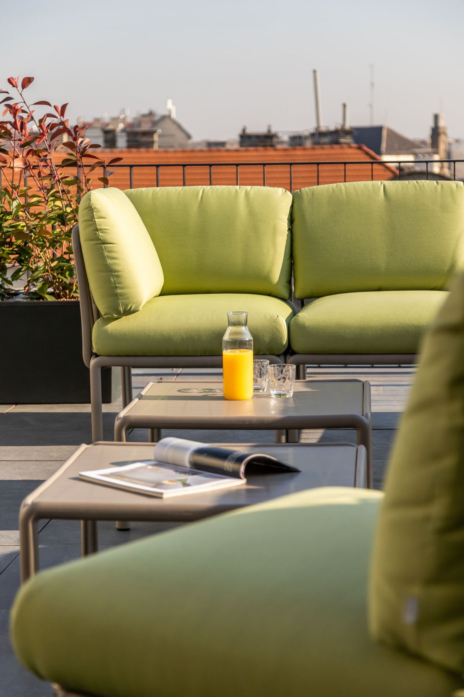 Stylish rooftop terrace with lime green cushioned sofas, small tables, orange juice bottle, and panoramic city views.