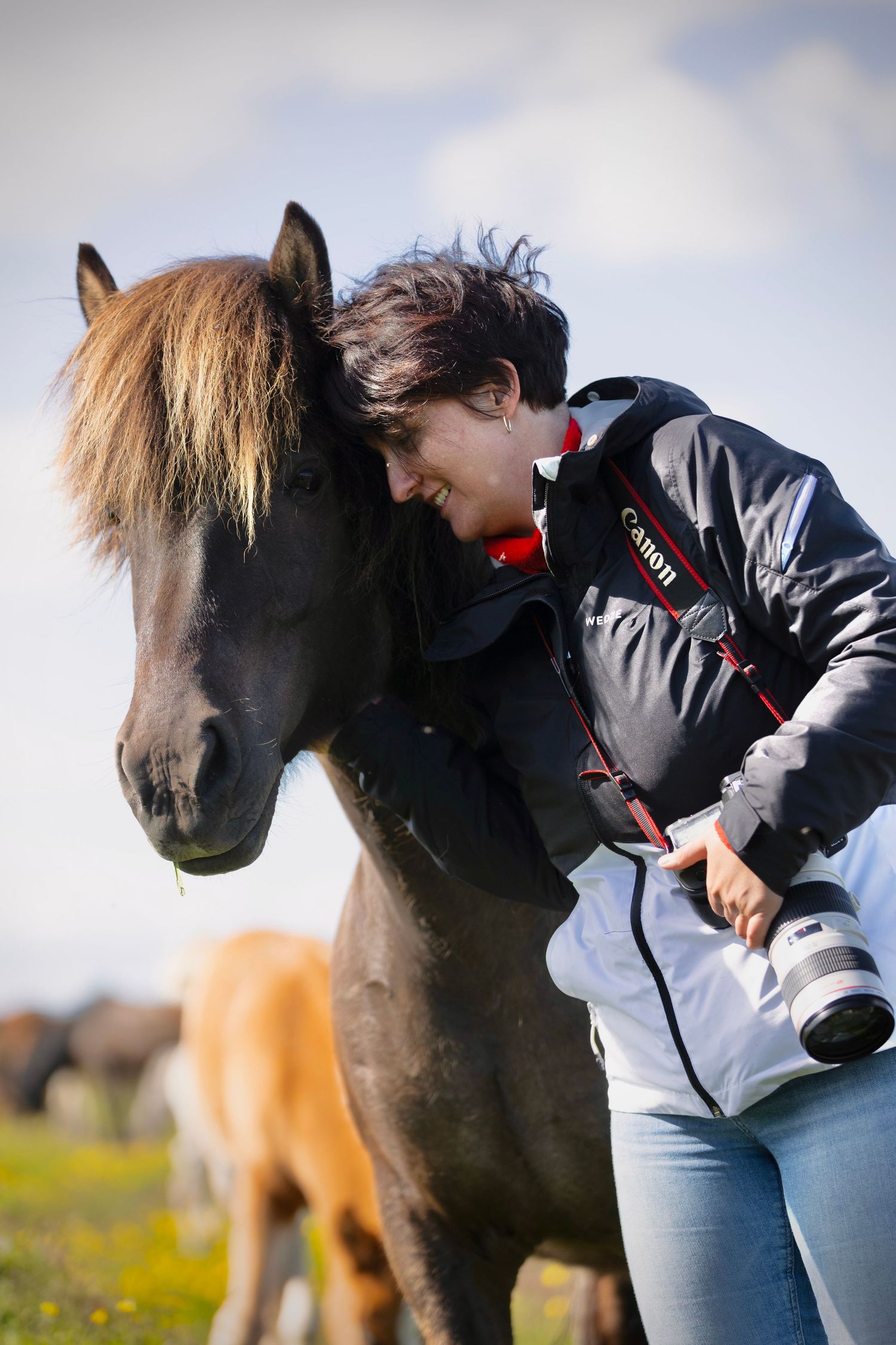 Female photographer with Canon camera smiling while leaning against an Icelandic horse outdoors.