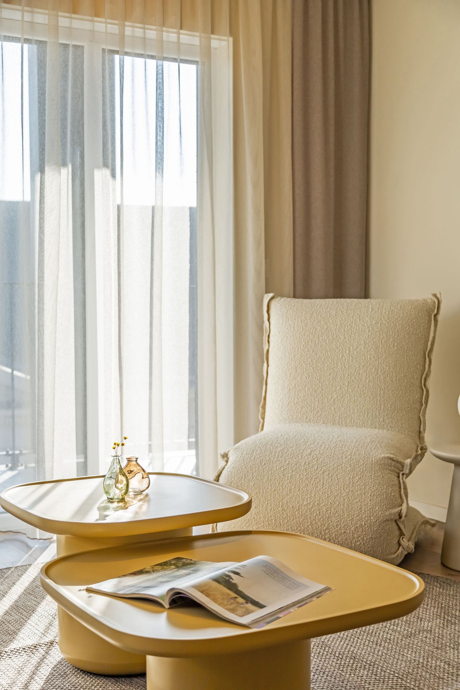 Bright hotel room corner with a white boucle armchair, yellow oval coffee tables, open magazine, and sheer curtains.