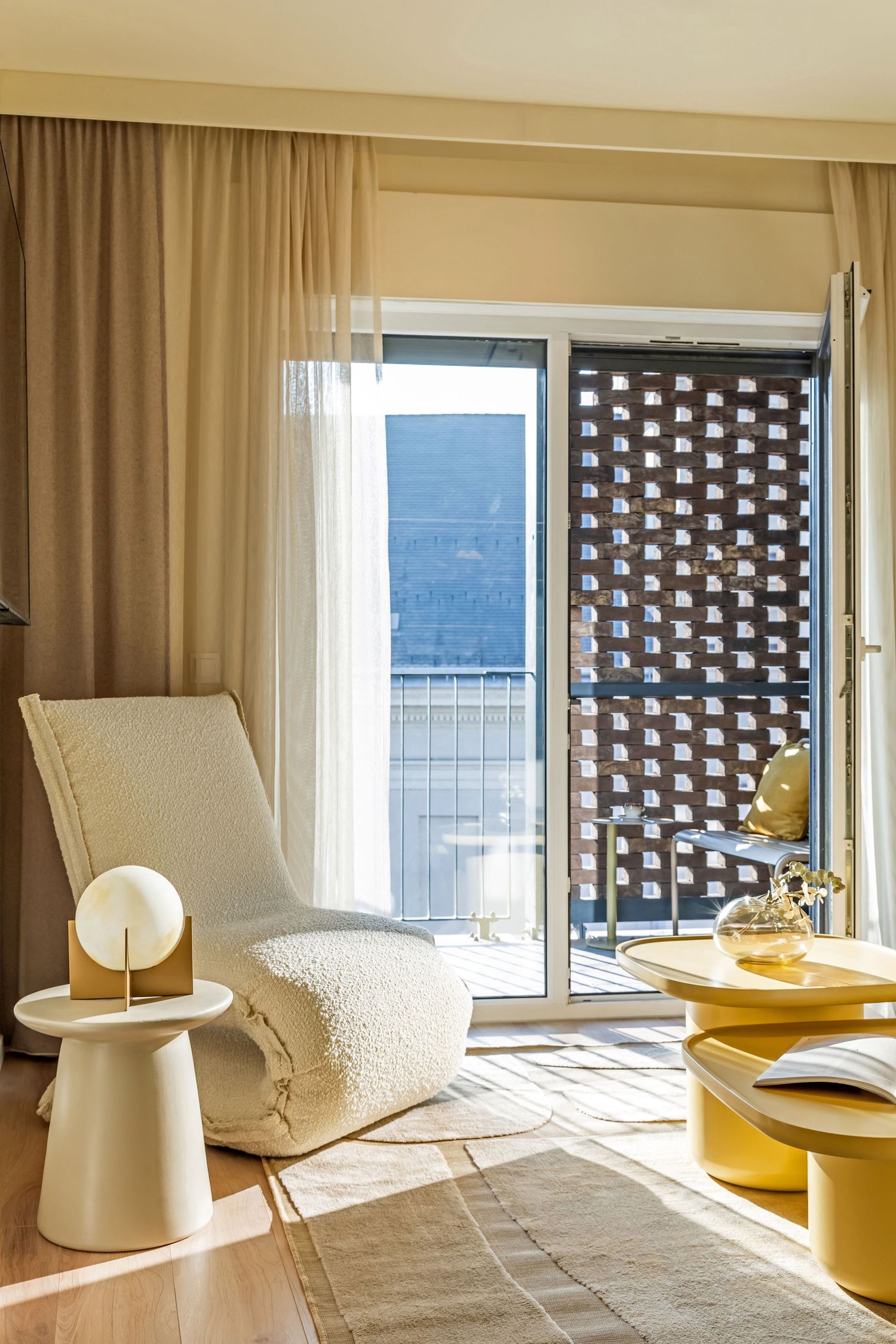 Sunny hotel room sitting area with white boucle lounge chair, round side table, yellow coffee table, and balcony view.