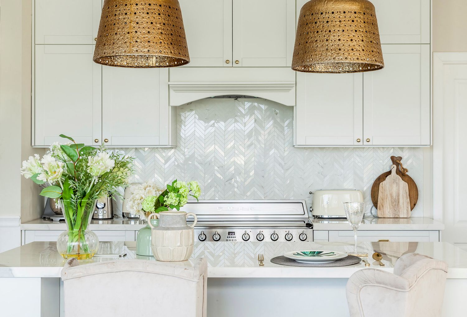 Elegant white kitchen featuring brass basket pendant lights and a herringbone marble tile backsplash.