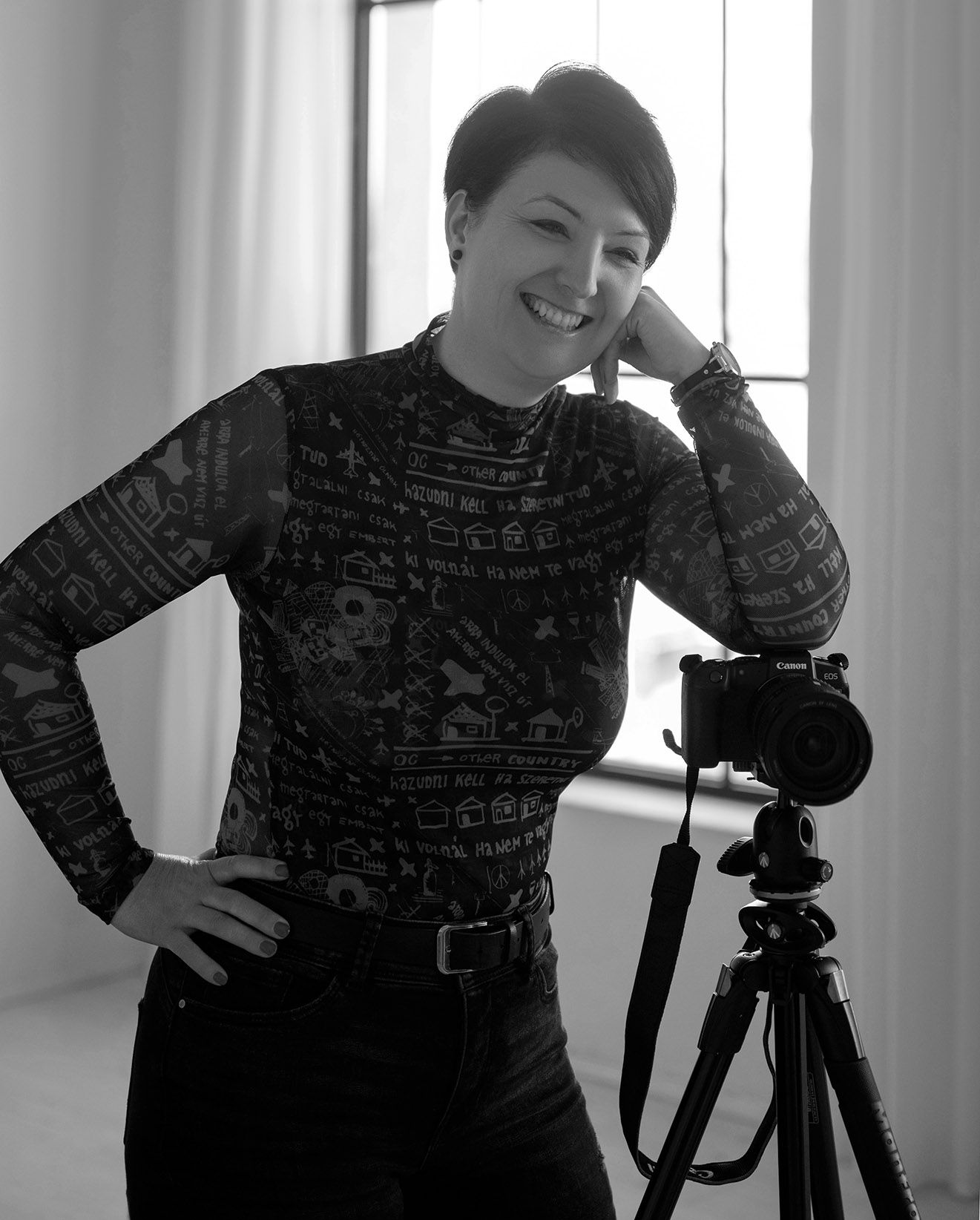 Black and white portrait of smiling woman with short hair standing next to camera on tripod.