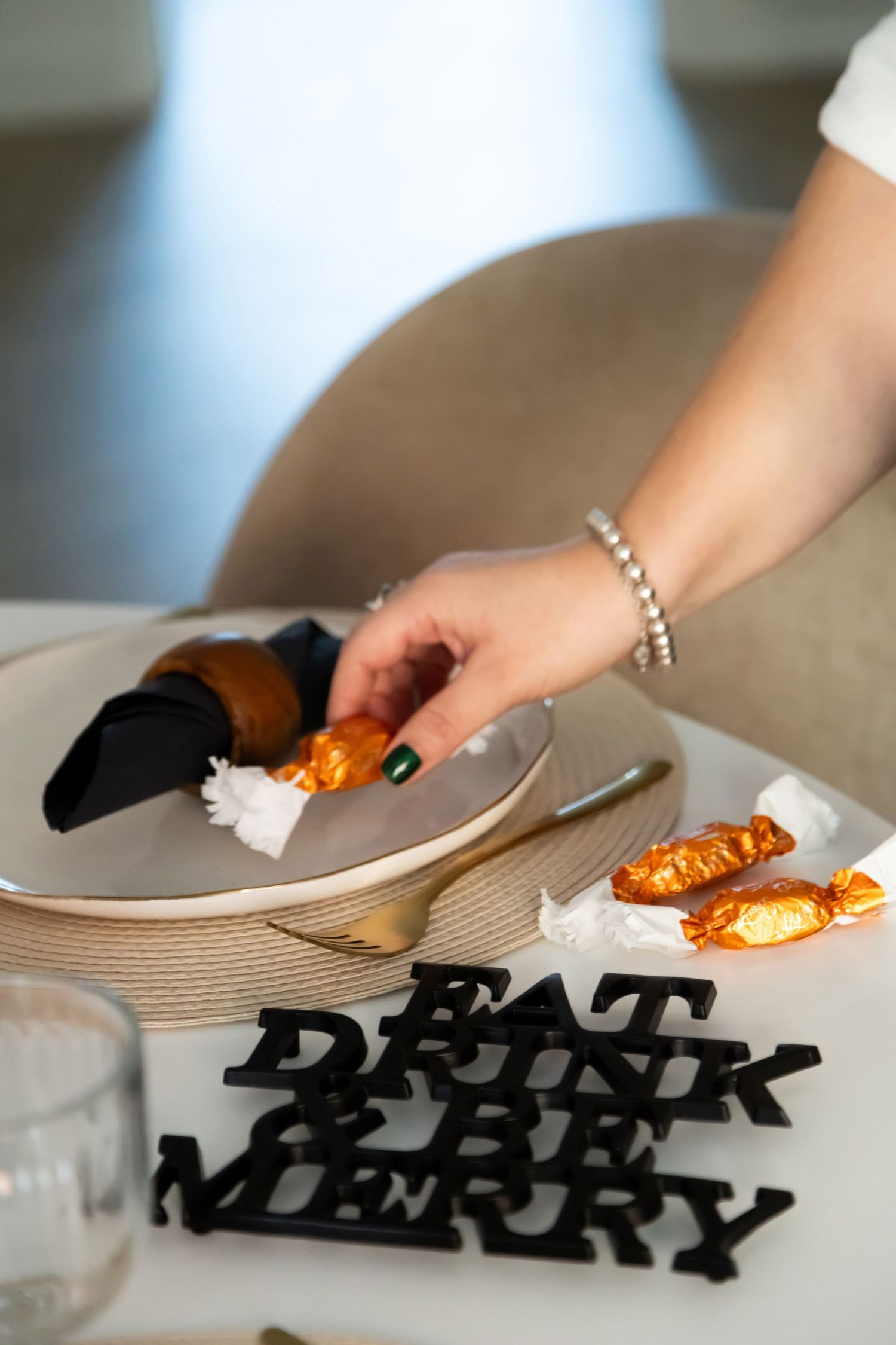 Close-up of a hand with green nails placing candy on a plate beside Eat Drink Be Merry holiday decor.