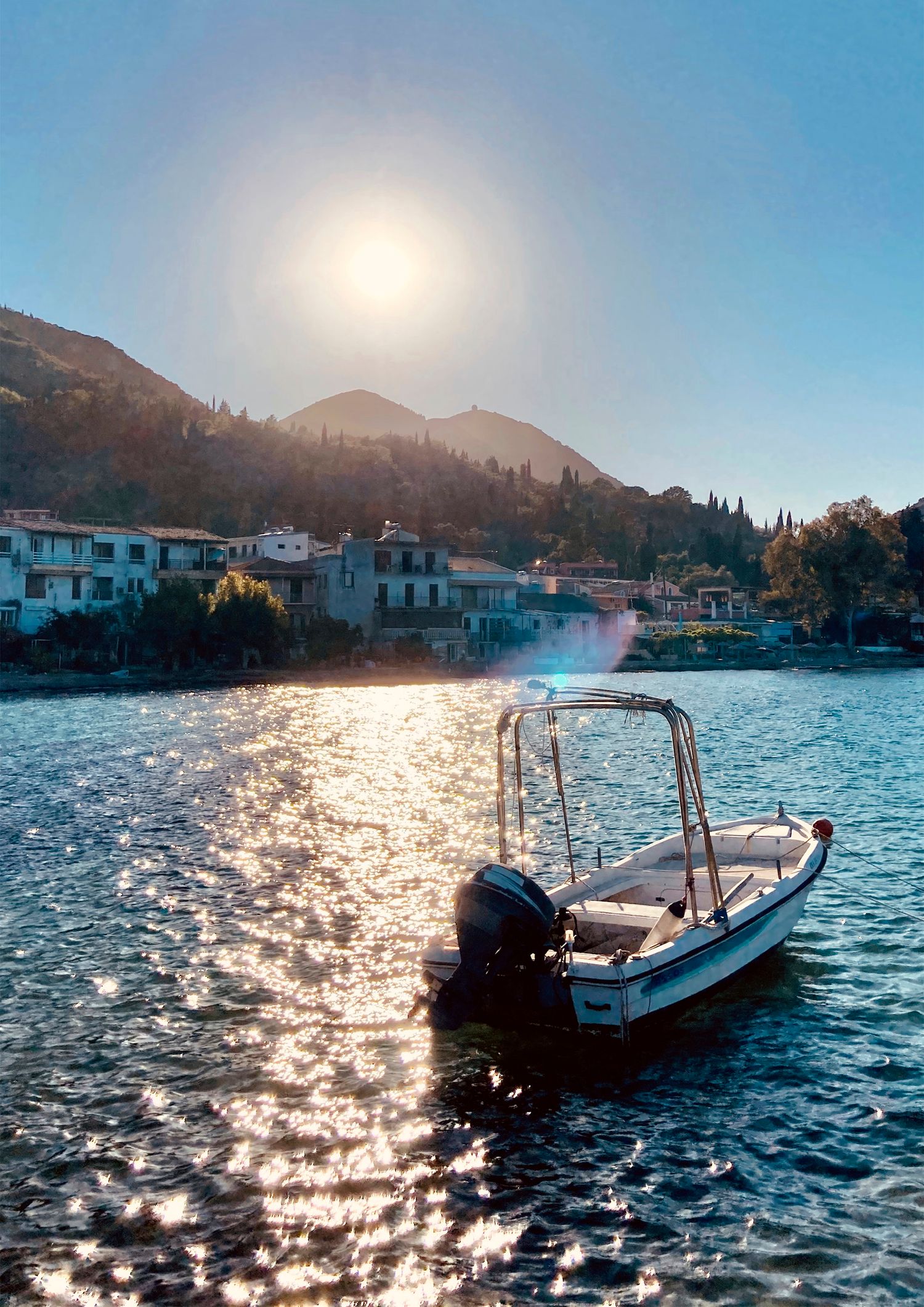A small motorboat anchored in a sunlit Mediterranean bay with a hillside village and mountains in the background.