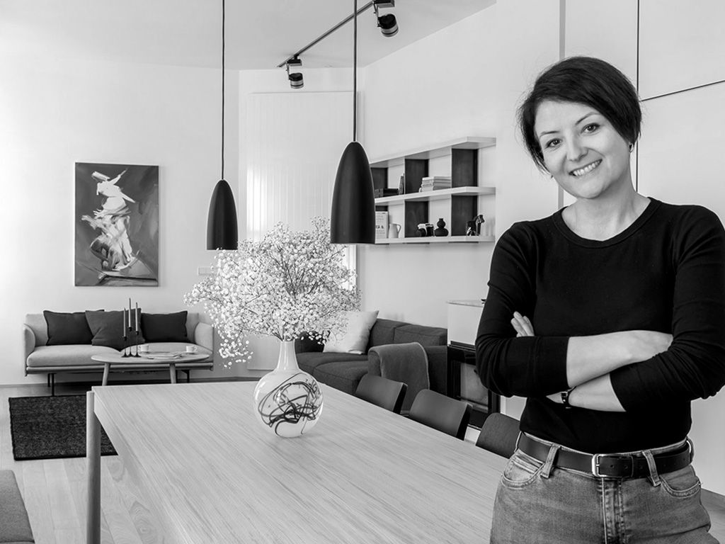 Black and white portrait of a smiling woman with arms crossed beside a dining table in a modern interior.
