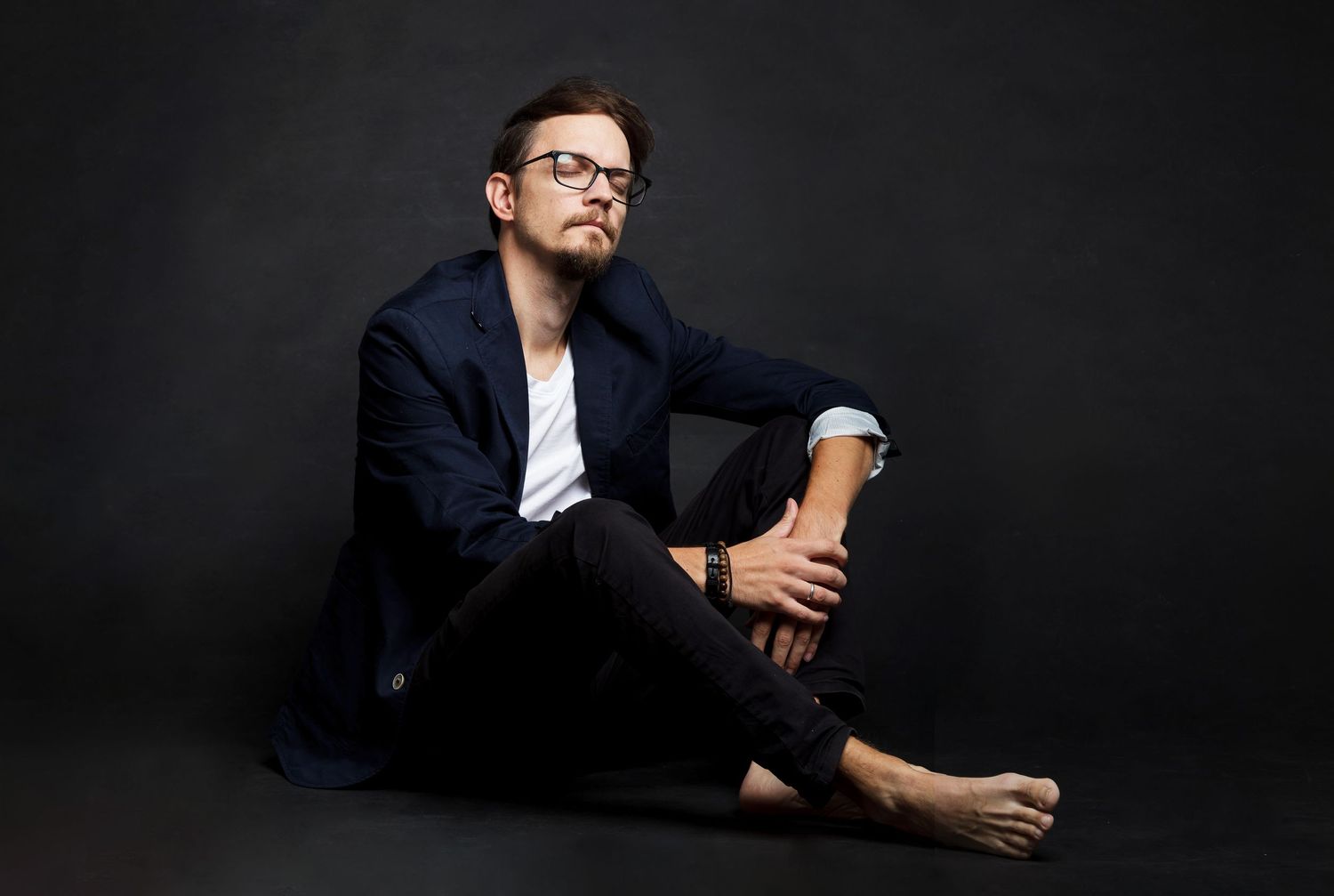 Stylish man in navy blazer and glasses sitting barefoot against dark studio background.