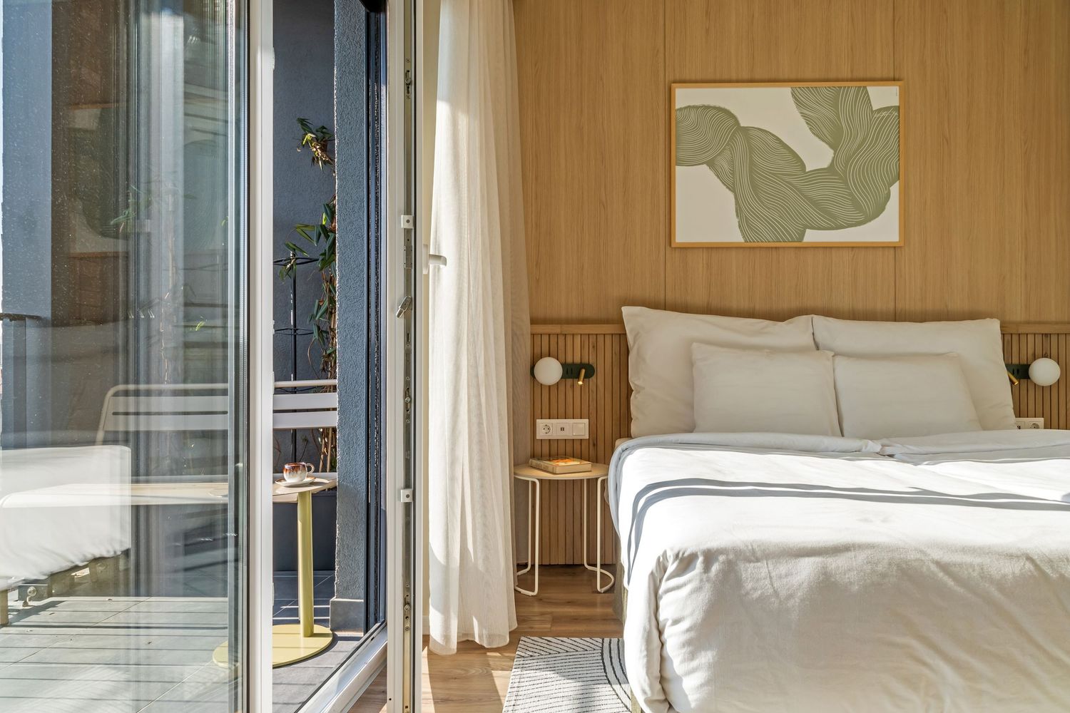 Minimalist hotel bedroom with wooden wall paneling, white bedding, abstract wall art, and glass balcony doors in warm light.
