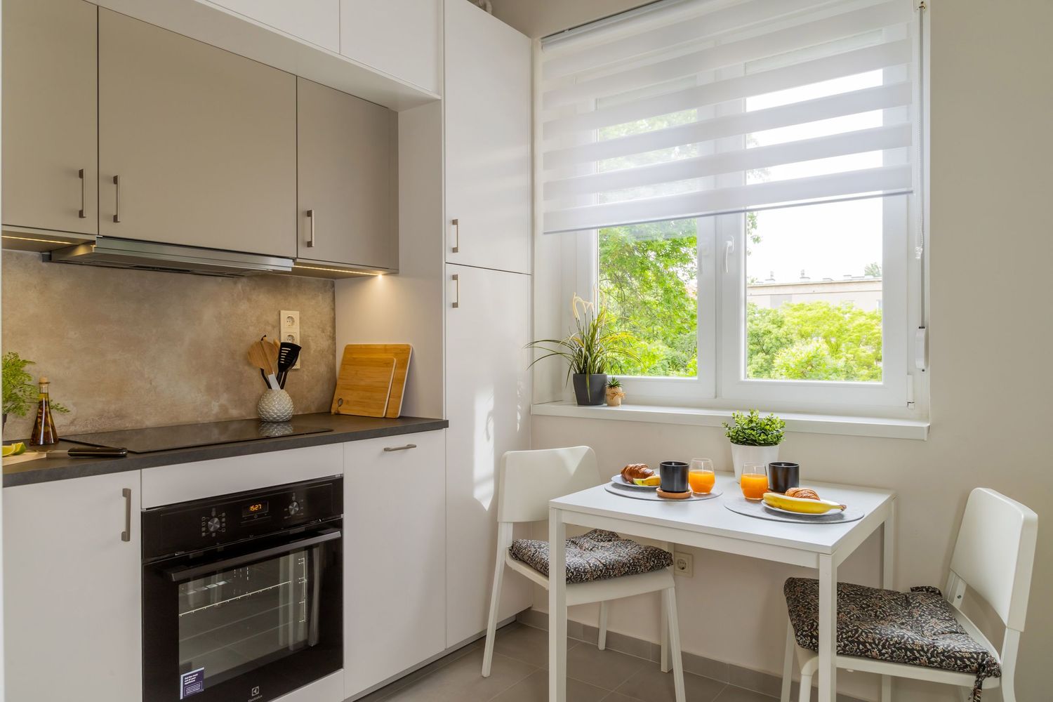 Compact modern kitchen with white cabinets, black oven, small dining table, and bright window with green view.