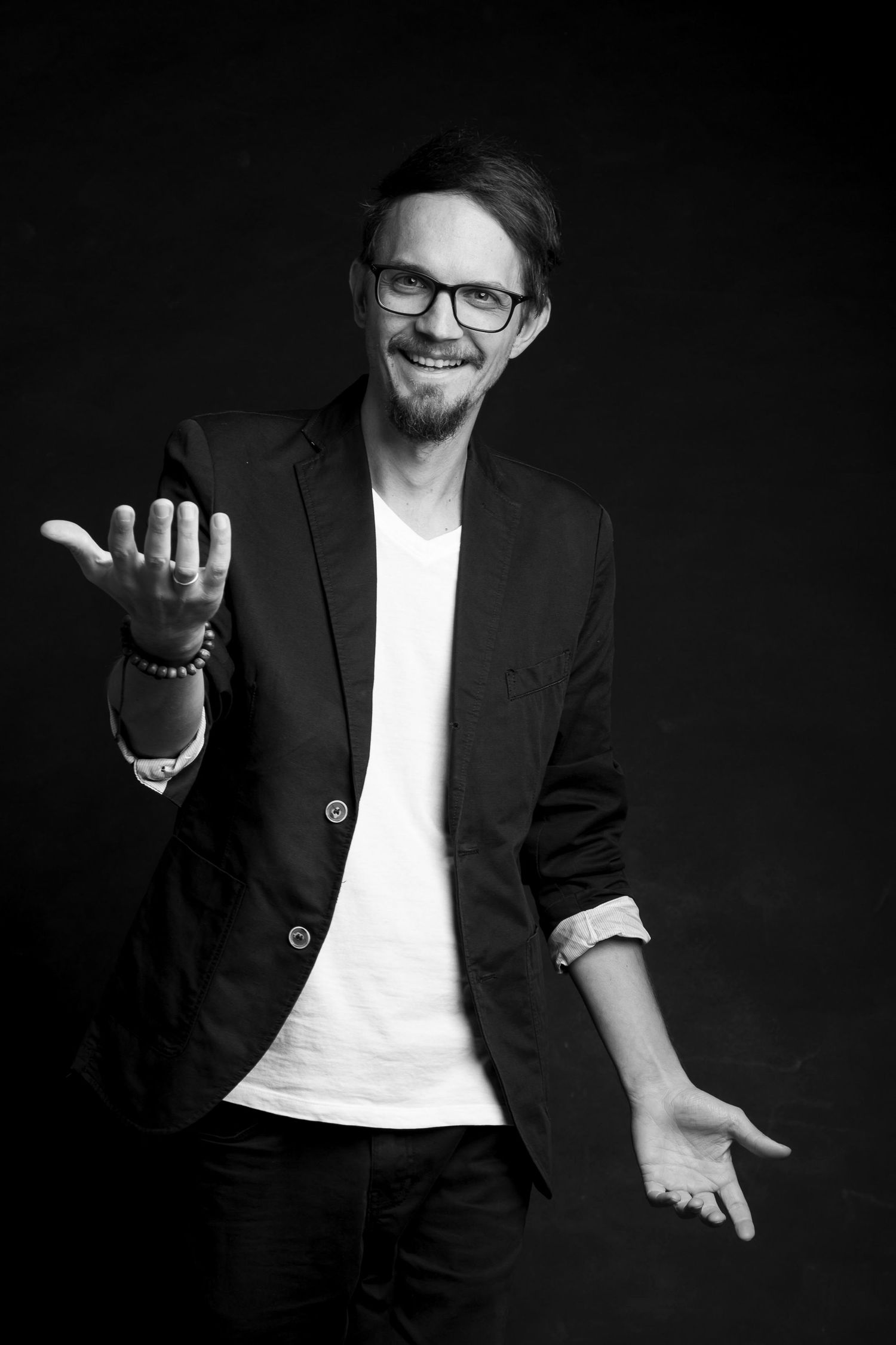Black and white portrait of smiling man in black blazer and glasses with open hand gestures.
