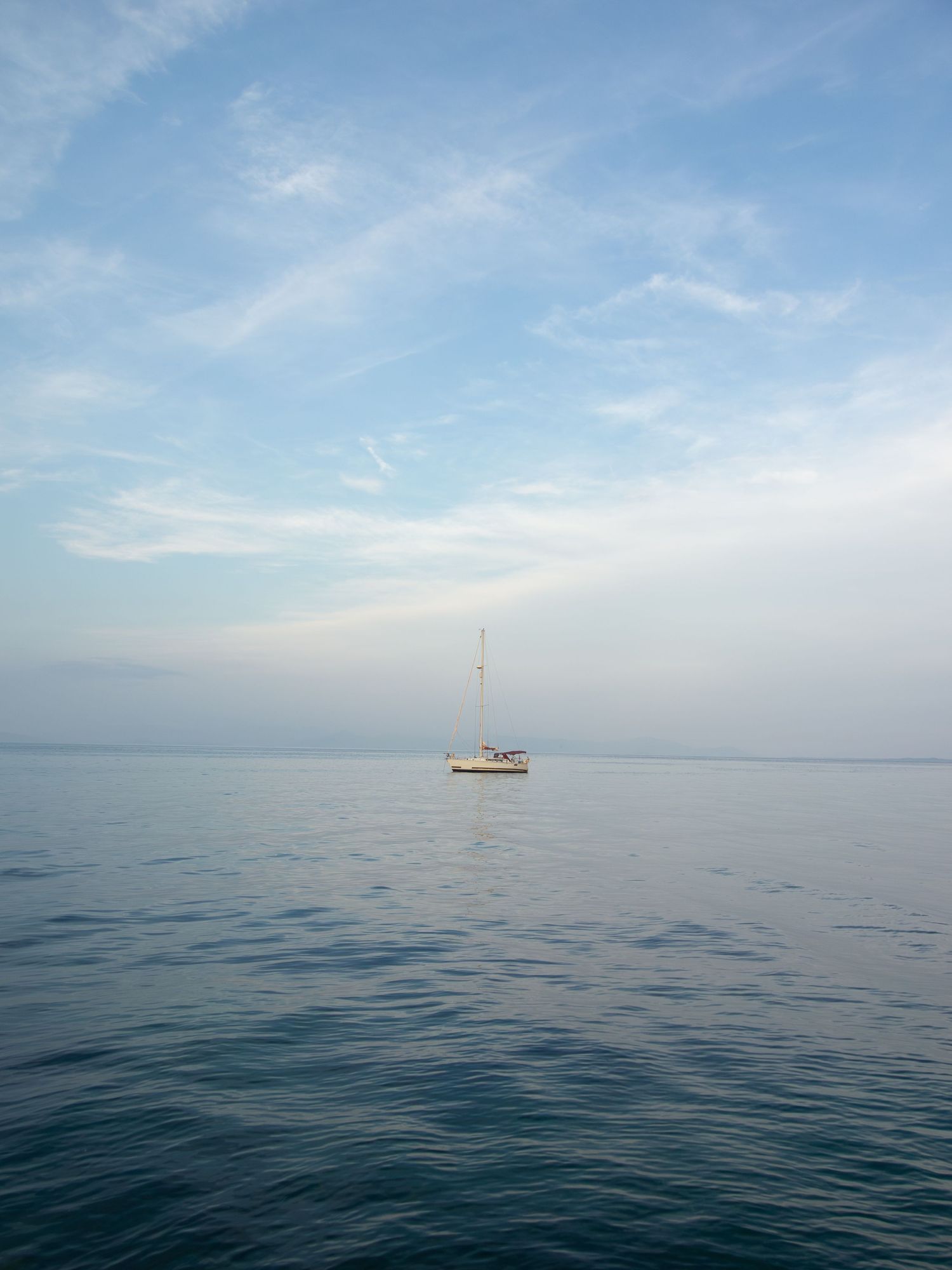 A lone sailboat floating on calm blue waters under a partly cloudy sky on a peaceful open sea.
