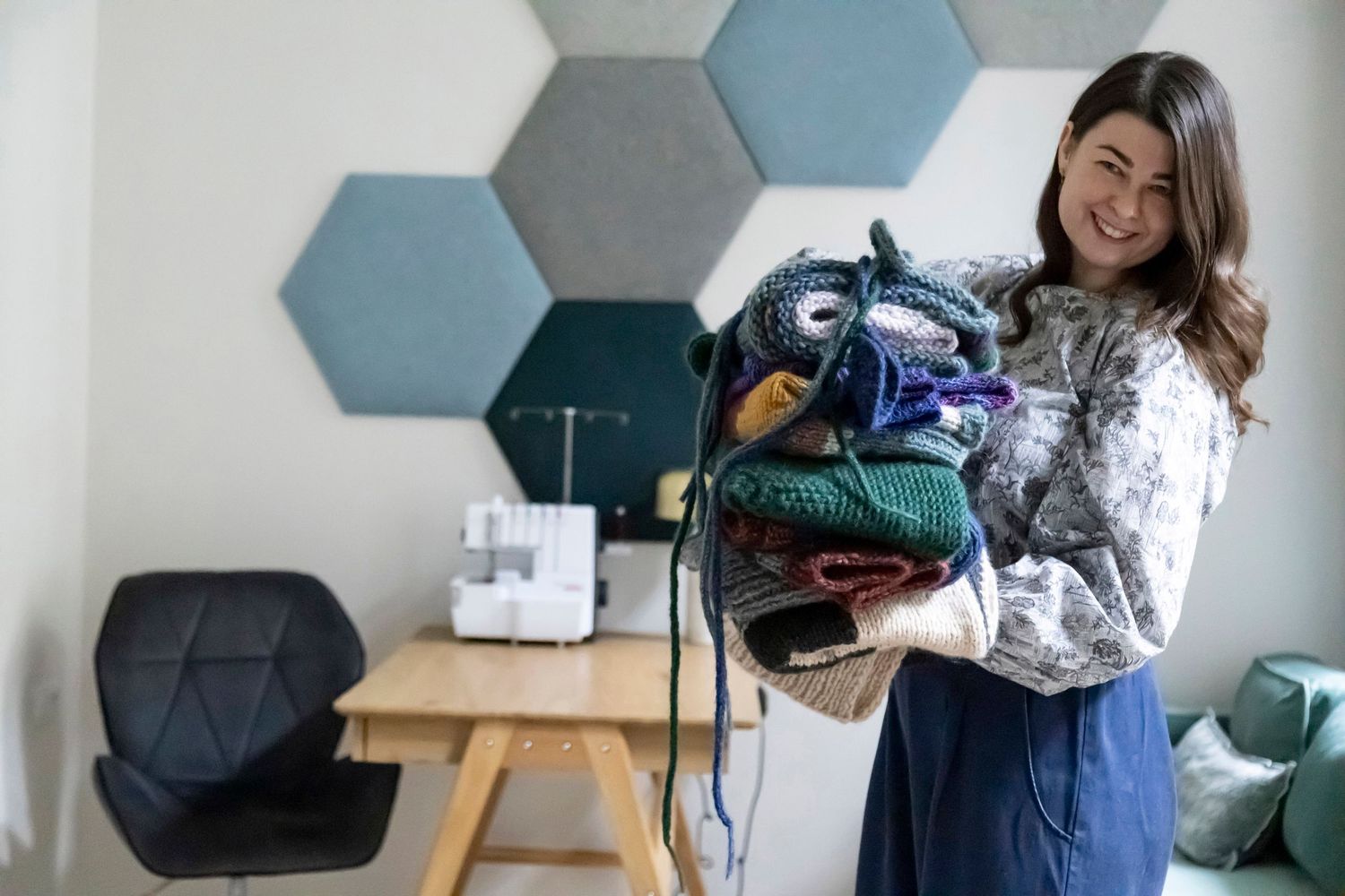 Smiling woman holding a large pile of colorful knitted items in a modern craft studio.