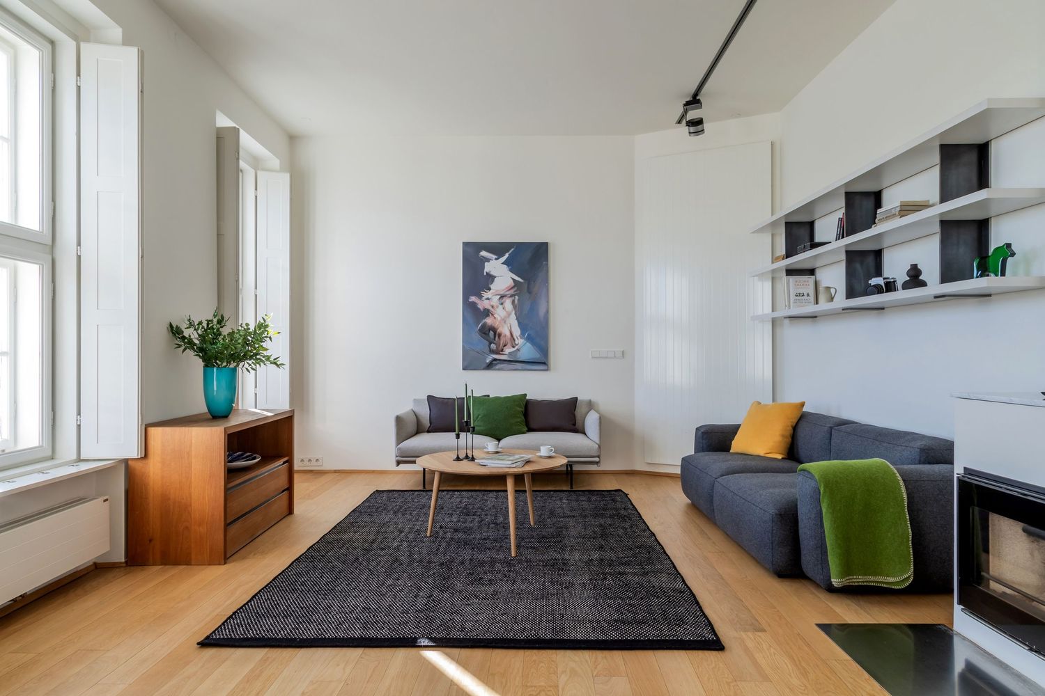 Minimalist living room with a blue sofa, dark rug, wooden floors, wall shelving, and a colorful abstract painting.