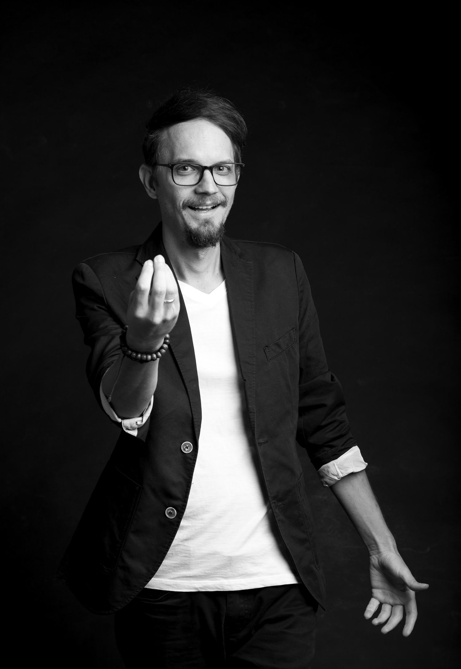 Black and white studio portrait of smiling man in blazer and glasses gesturing toward camera with hand.