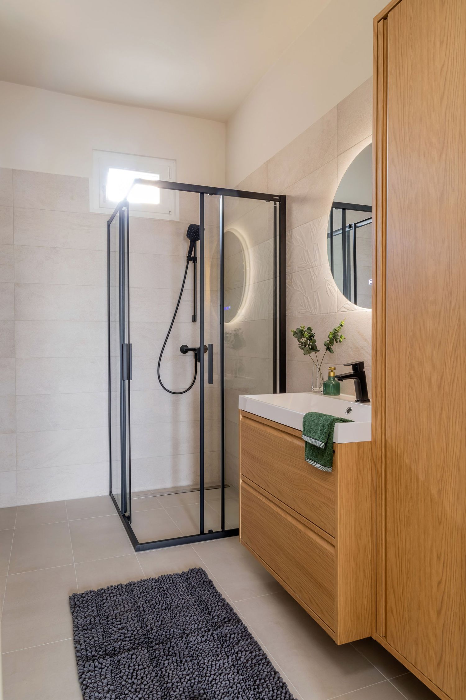 Modern bathroom featuring black-framed glass shower, oak wood vanity, round LED mirror, and gray textured bath mat.