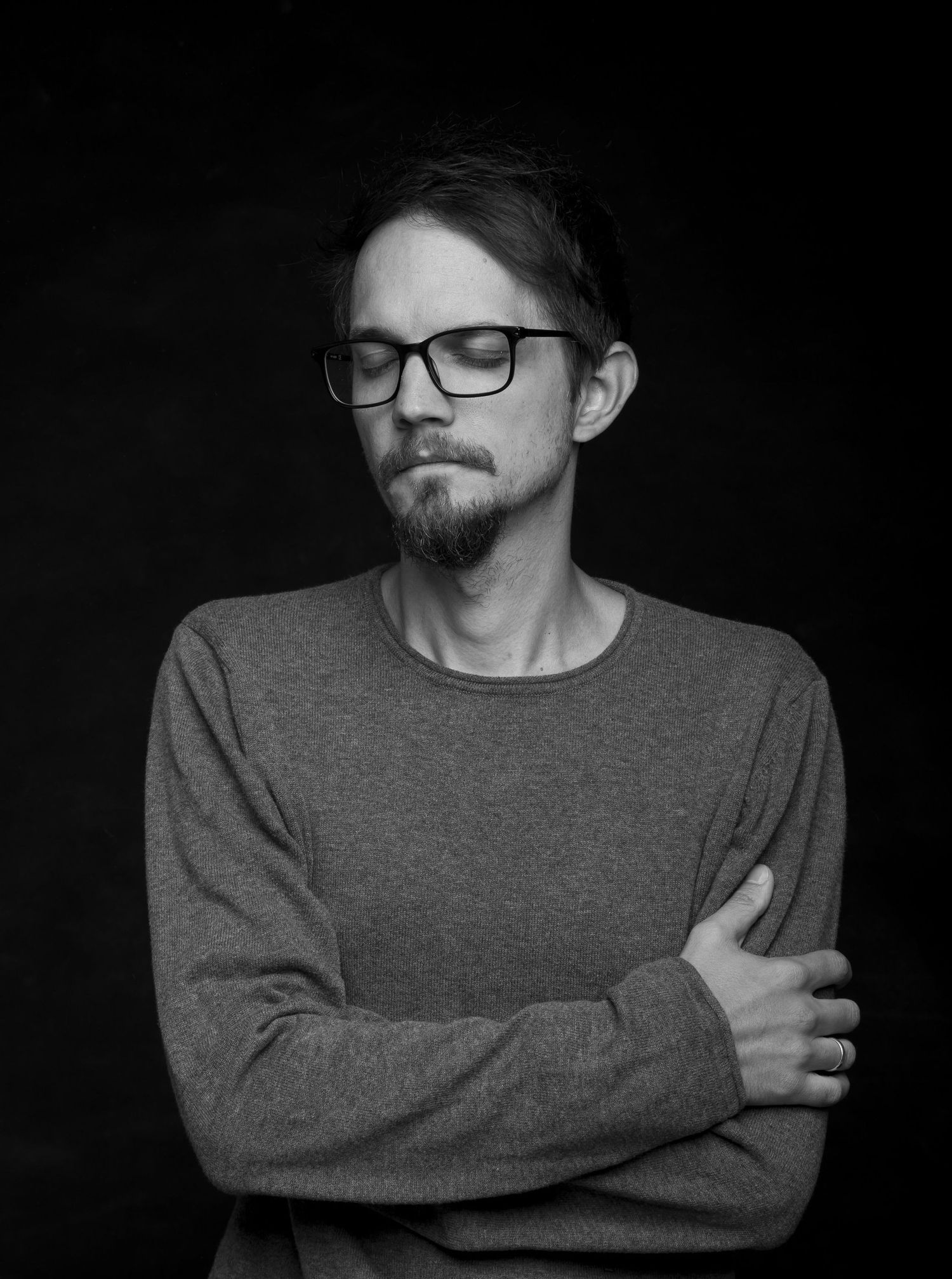 Black and white portrait of man in gray sweater and glasses with arms crossed and eyes closed.