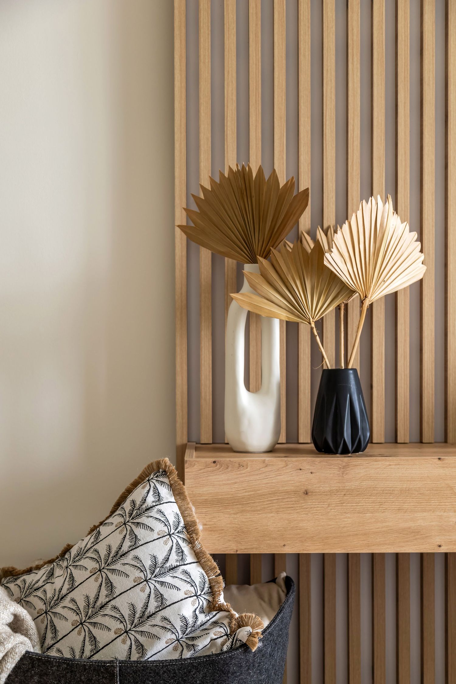 Decorative wooden shelf with dried palm leaf arrangements in white and black vases against a slatted wood wall.